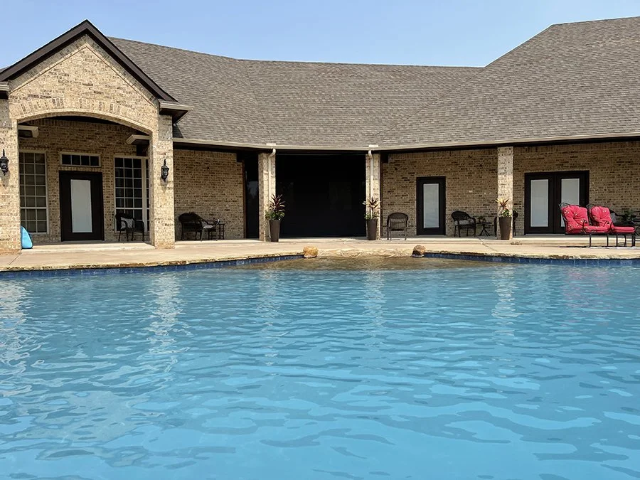Backyard pool area with brick house, patio chairs, potted plants, and poolside seating.
