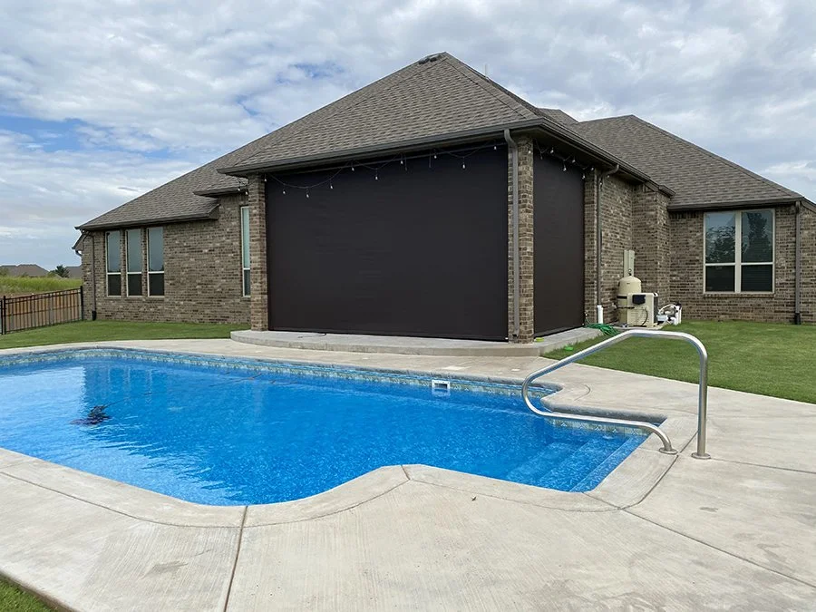 Backyard view of a house with a swimming pool and a large black screen or cover attached to the house, with pool stairs visible and a grassy area around the concrete patio.