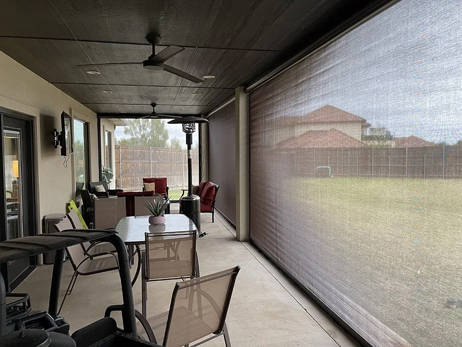Covered outdoor patio with chairs, a table with a potted plant, and a ceiling fan, overlooking a backyard with a fenced yard and houses in the distance.
