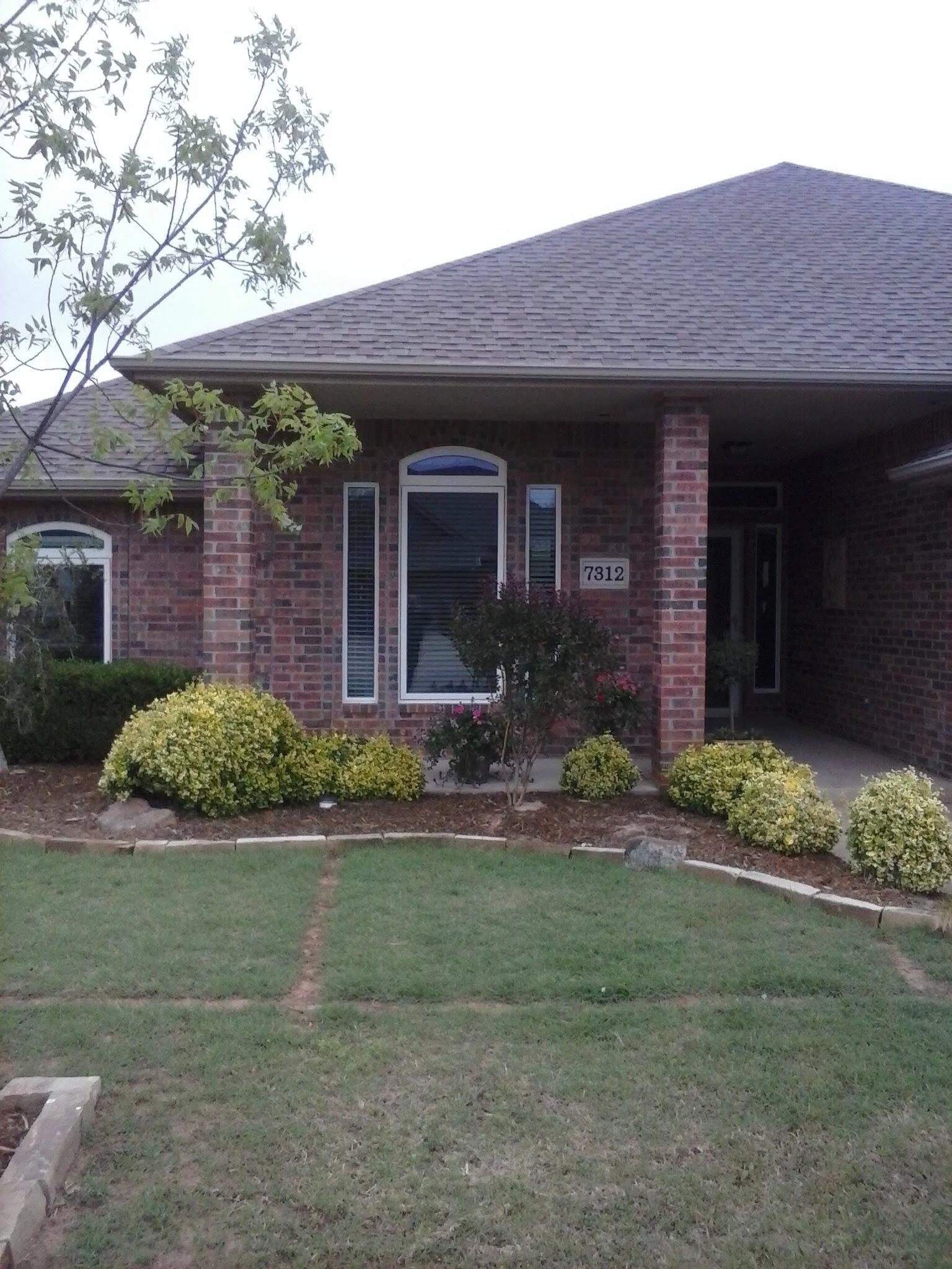 A brick house with a gable roof, a large front window, and a porch. The house number is 7312. There are green bushes, small plants, and grass in the front yard.