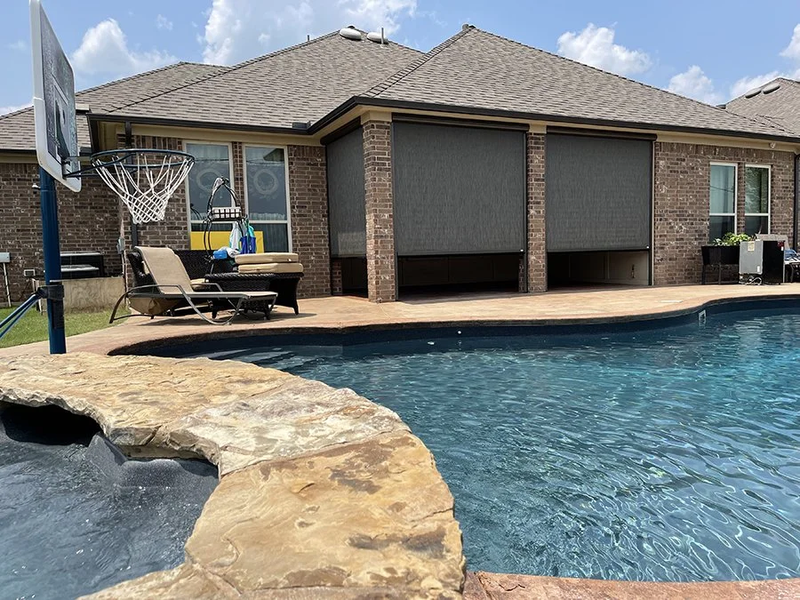 Backyard scene featuring a swimming pool with a hot tub, outdoor lounge chairs, a basketball hoop, and a brick house with screened porch windows and closed shades.