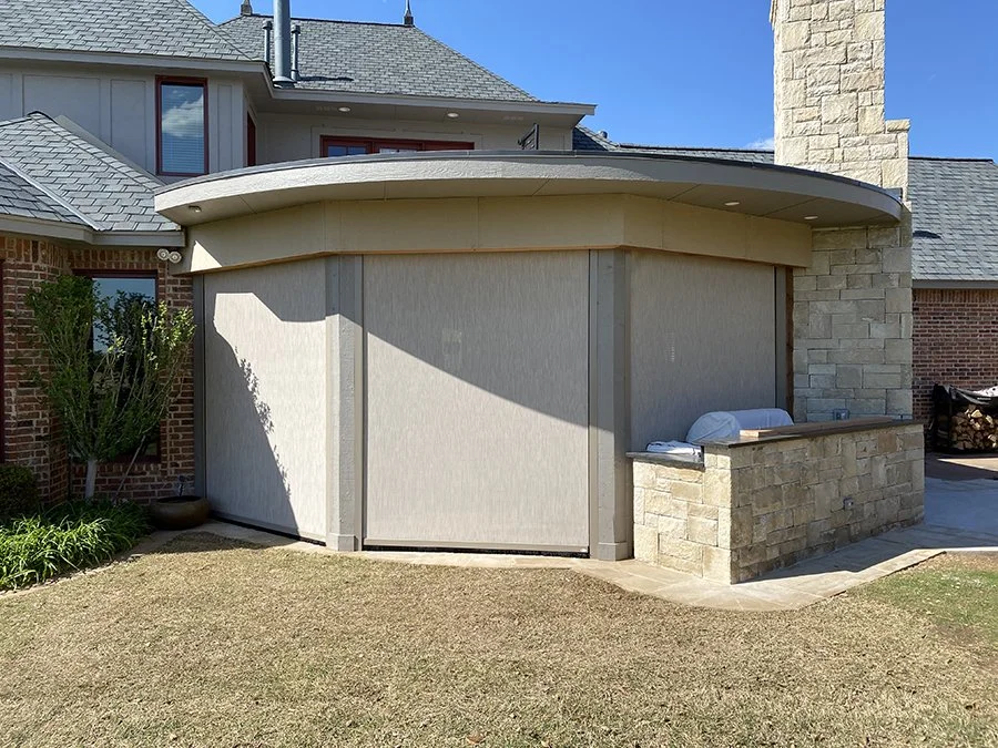 A house with a curved, light-colored wall and roof, brick walls, and a stone chimney. There is a small outdoor area with pillows and a stone wall, and a lawn area in the foreground.