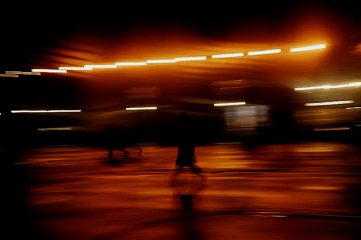 Blurred night scene with bright lights and a few people standing on a wet surface.