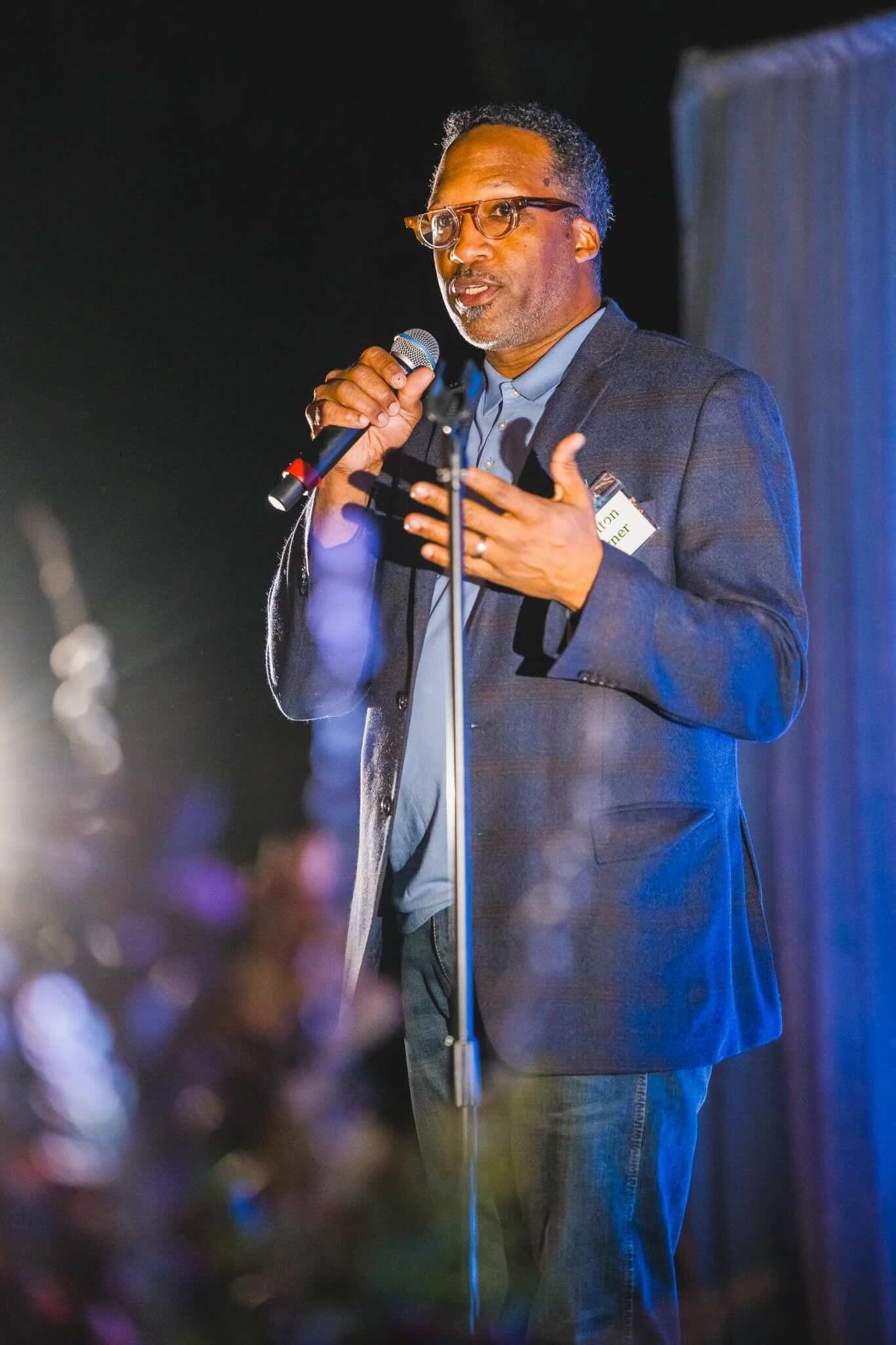 A man in glasses and a blazer speaking on stage at CFM's Friendsgiving event.