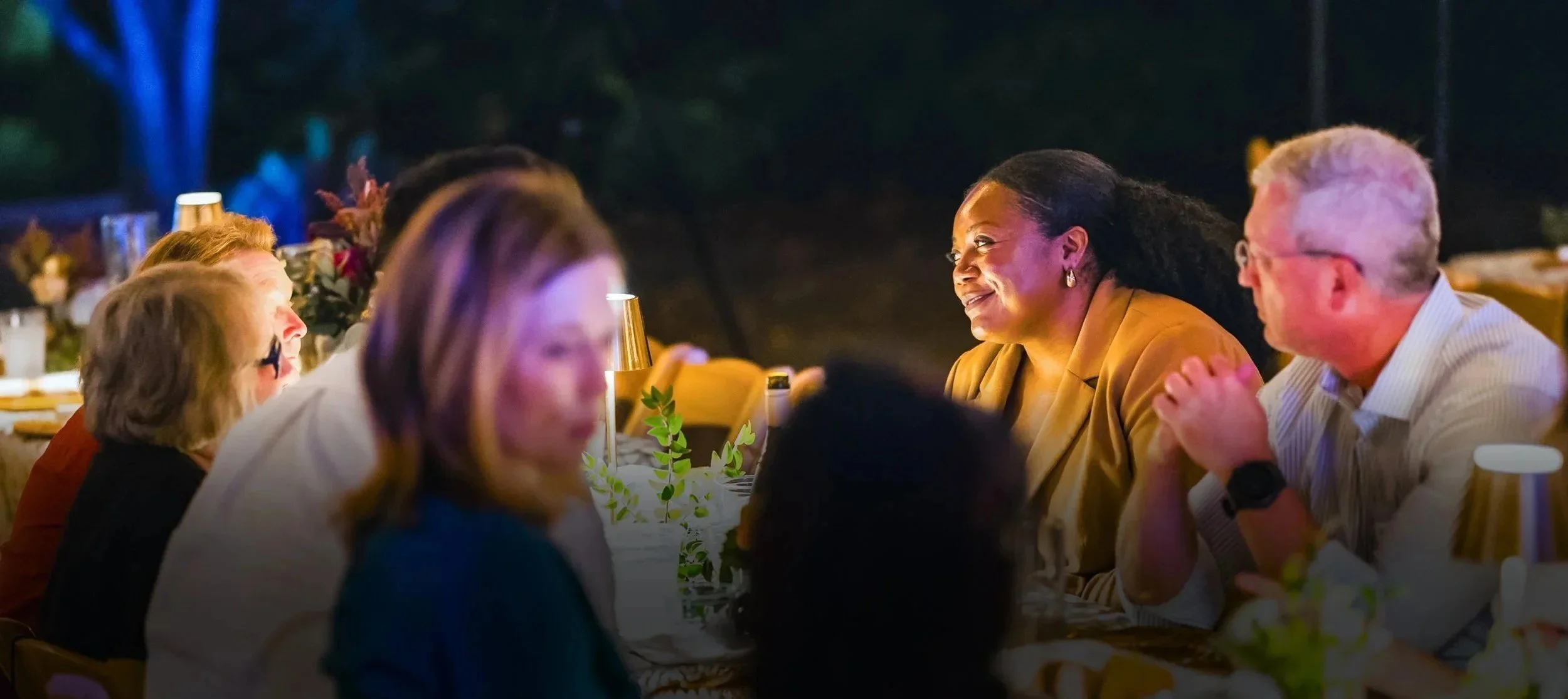 People sitting at a dinner table outdoors at CFM's Friendsgiving event, engaging in conversation and smiling, with candles and decorations on the table.