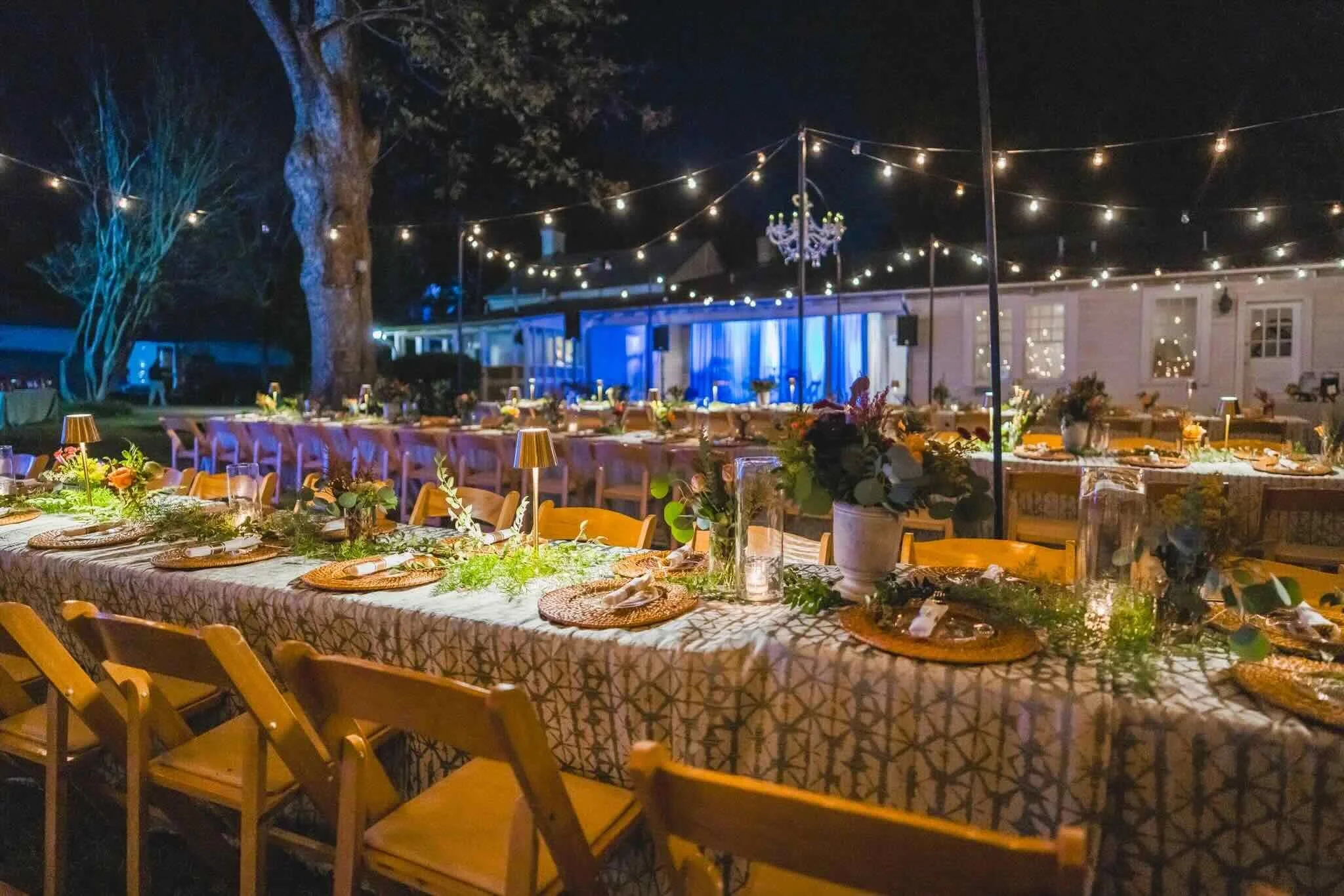 CFM's Friendsgiving event setup with long decorated tables, string lights, and a chandelier hanging from a pole, surrounded by trees and a nearby building illuminated with lights.