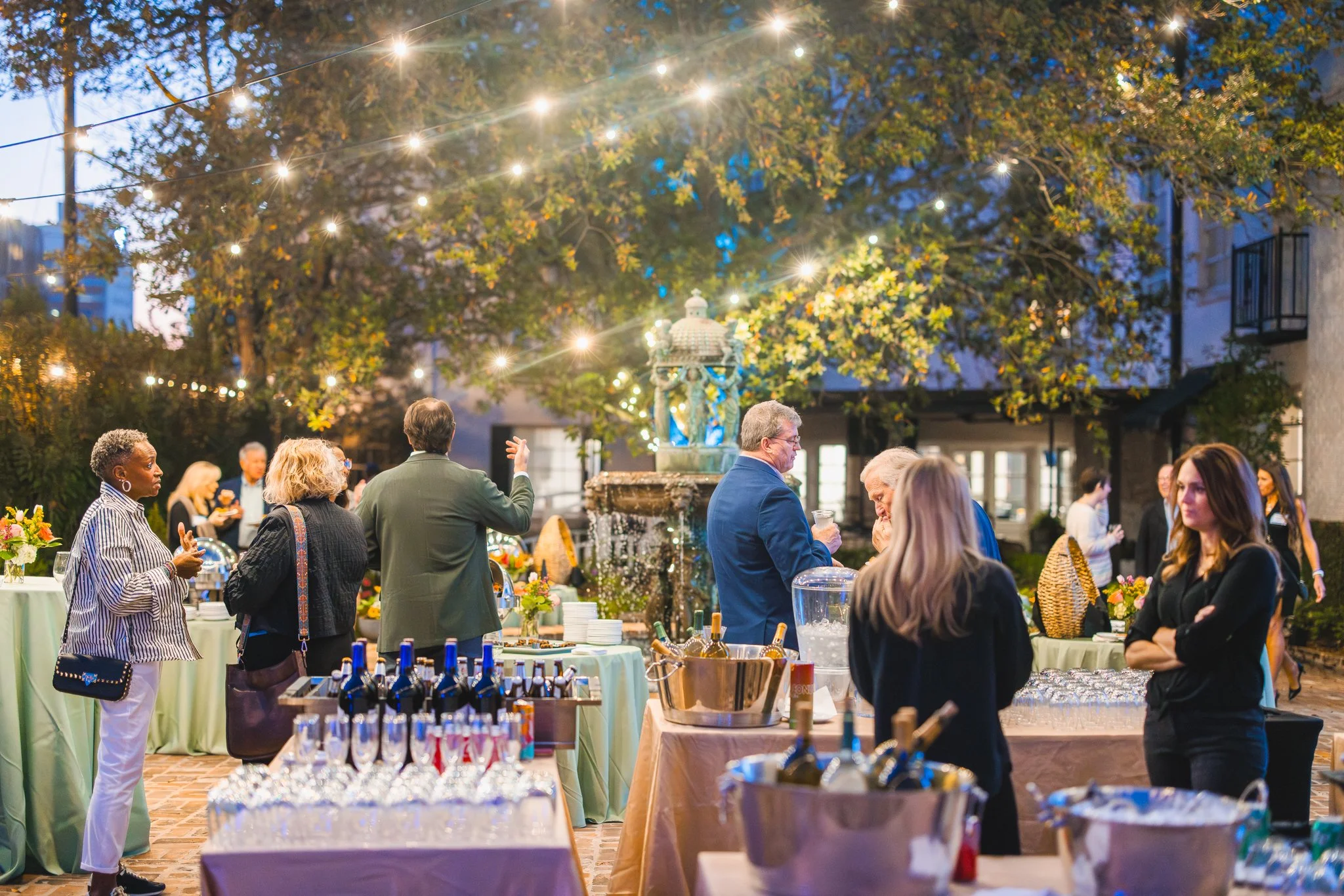A gathering of people around tables with food and drinks, under string lights hanging above, with trees and buildings in the background. CFM Giving Forward