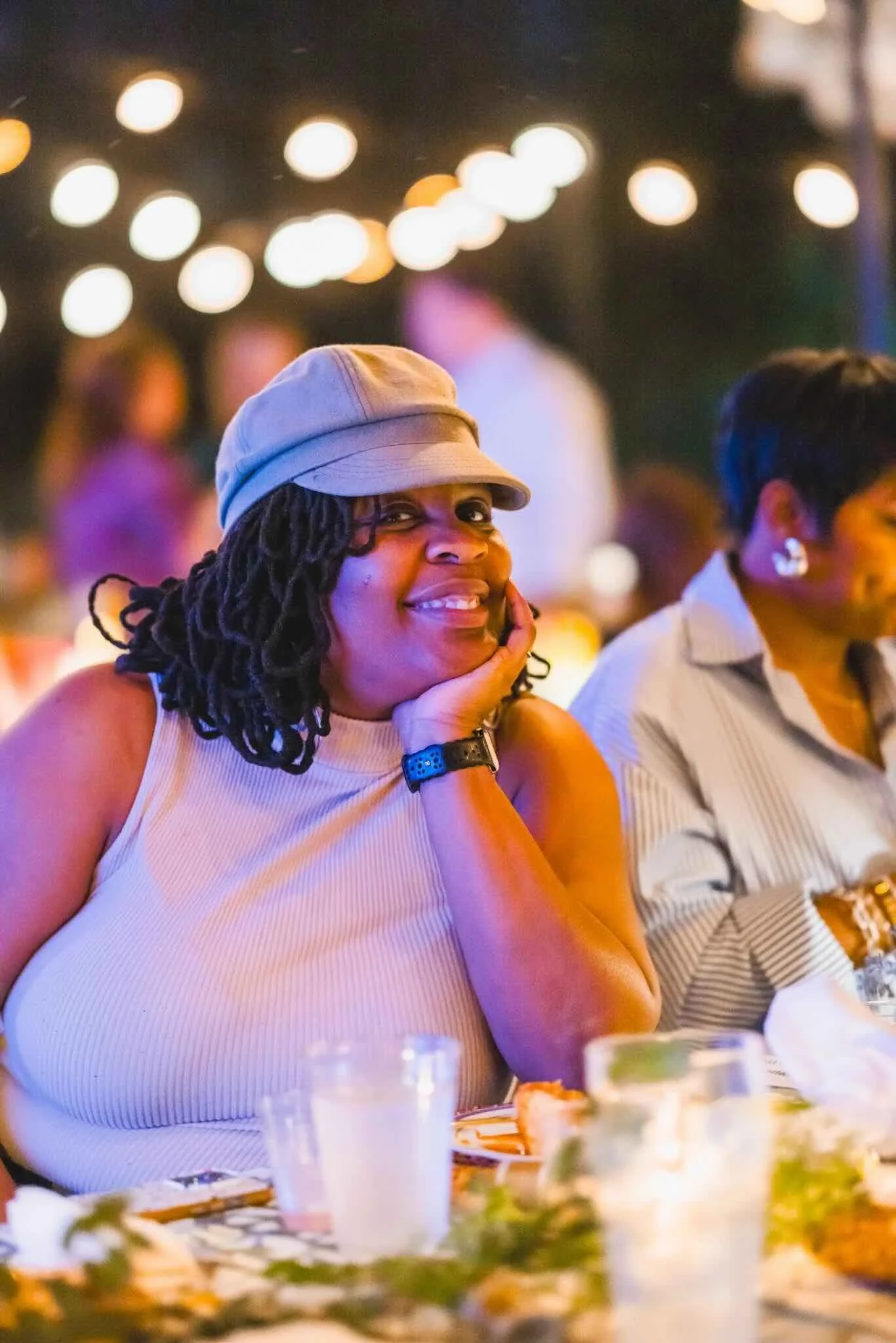 A woman with dreadlocks and a beige cap is smiling and resting her chin on her hand at a nighttime social gathering, with blurred lights and other people in the background.