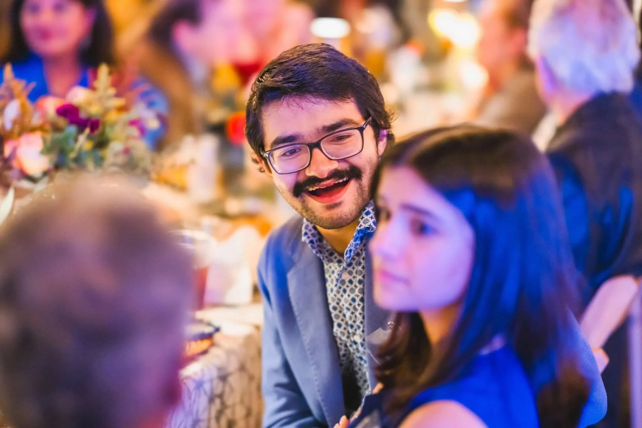 A man wearing glasses and a patterned shirt, smiling and engaging in conversation, colorful lights and floral decorations in the background.