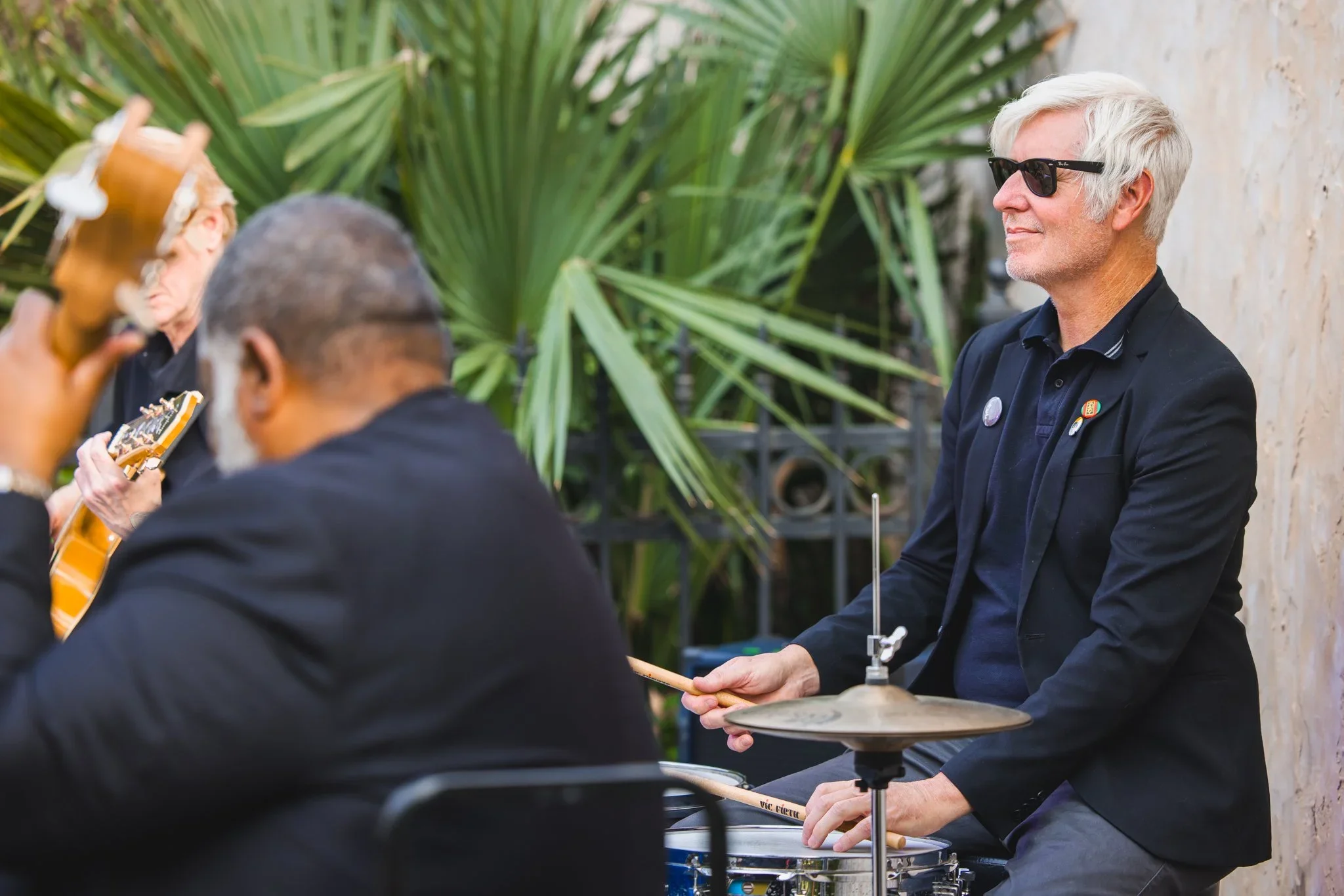 A man with white hair, wearing sunglasses and a black jacket, sitting outdoors near green plants and a stone wall, holding a drumstick and playing a drum.