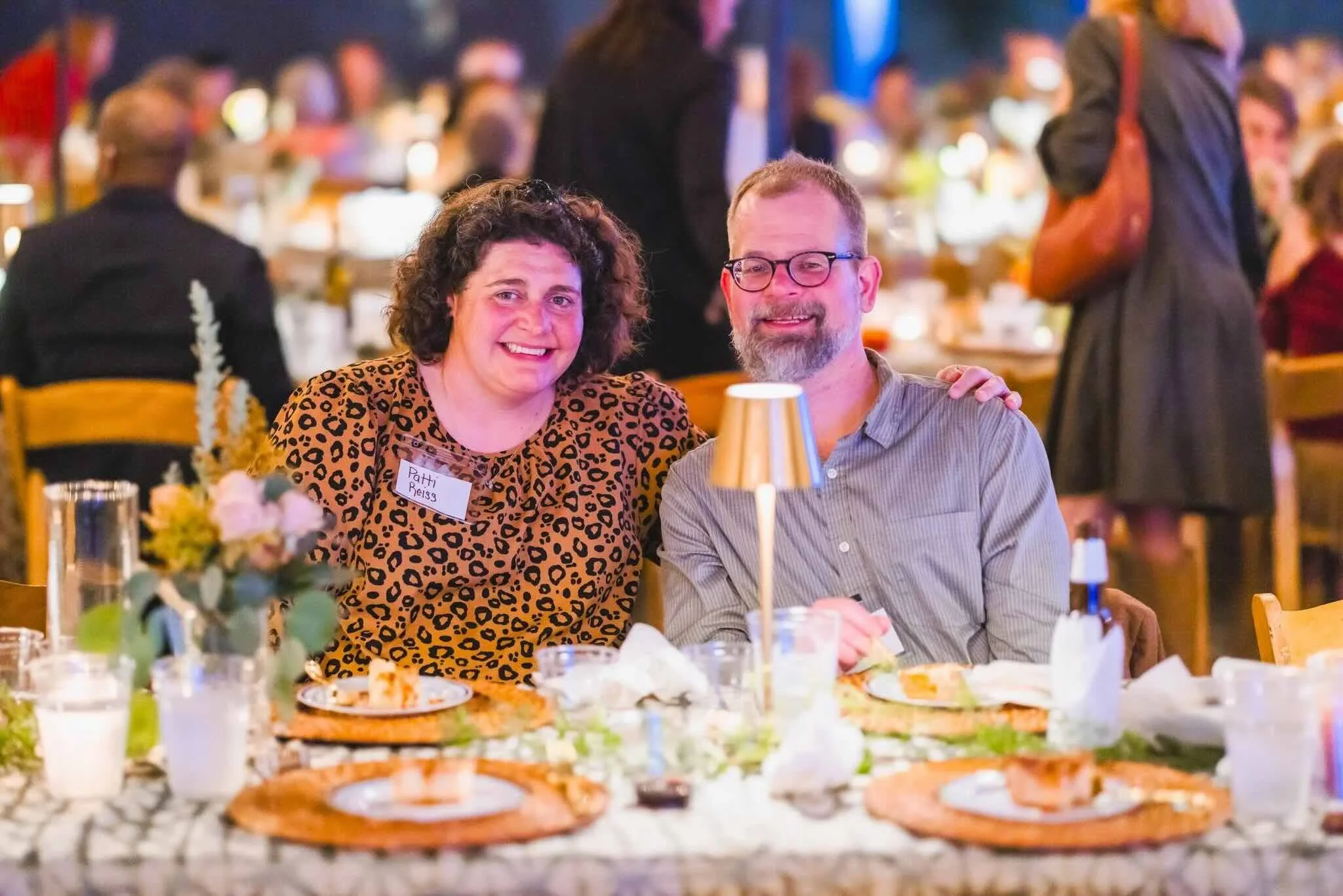 A smiling woman with curly hair wearing a leopard print top and a man with glasses and a beard wearing a striped shirt seated at a dinner table in a lively restaurant, surrounded by food and drinks.