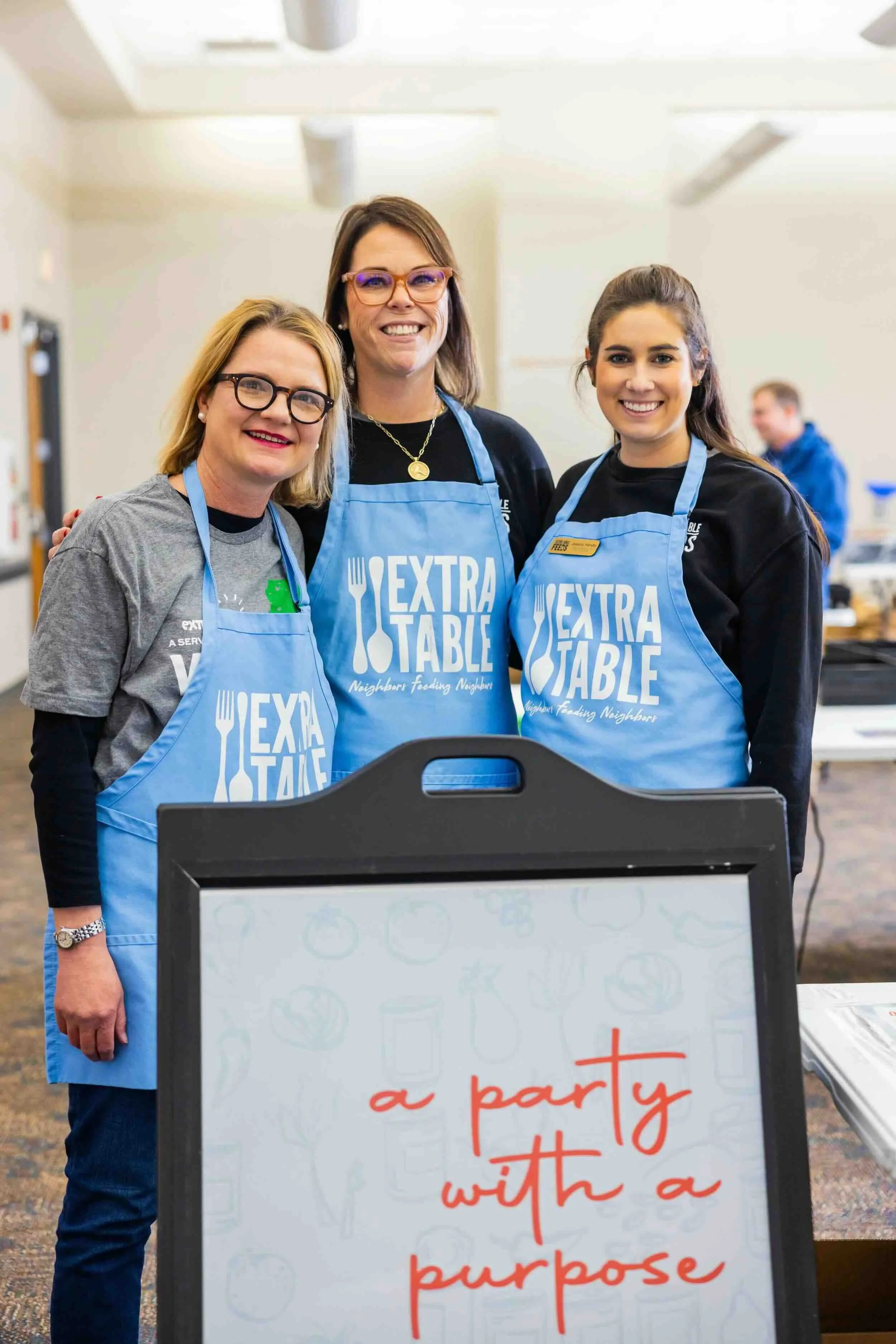 Three women in Extra Table aprons stand around a sign. | Extra Table Packing Party | Community Foundation for Mississippi Grants