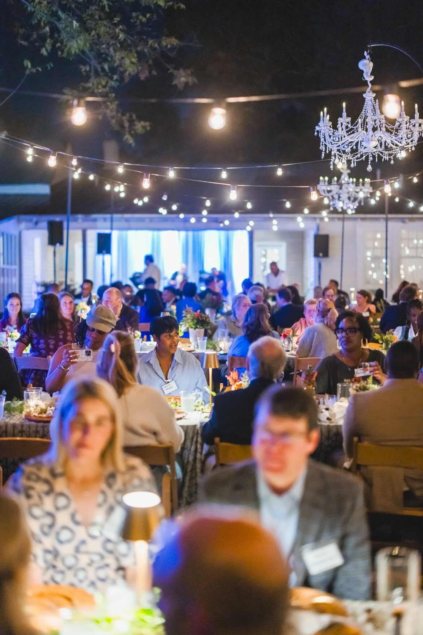 A gathering of people at CFM's Friendsgiving event with string lights and chandeliers hanging overhead. The scene features multiple tables with guests dining and engaging in conversations indoors or under a covered outdoor space.