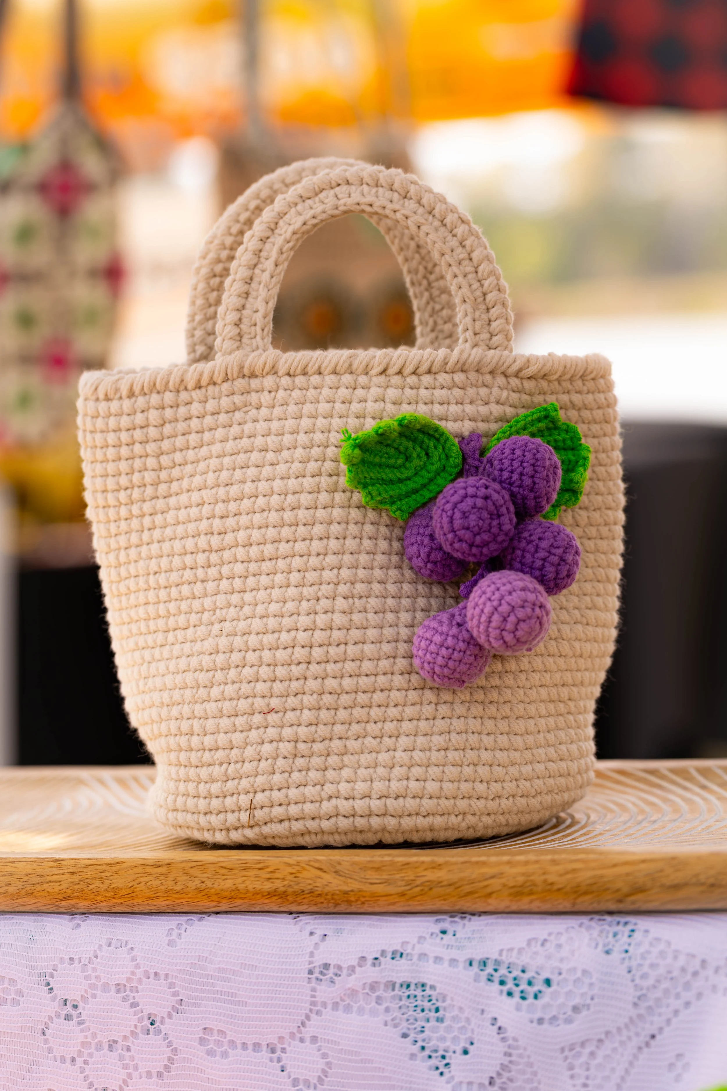 A cream-colored crocheted handbag with a purple and green crocheted grape cluster decoration, placed on a wooden surface.