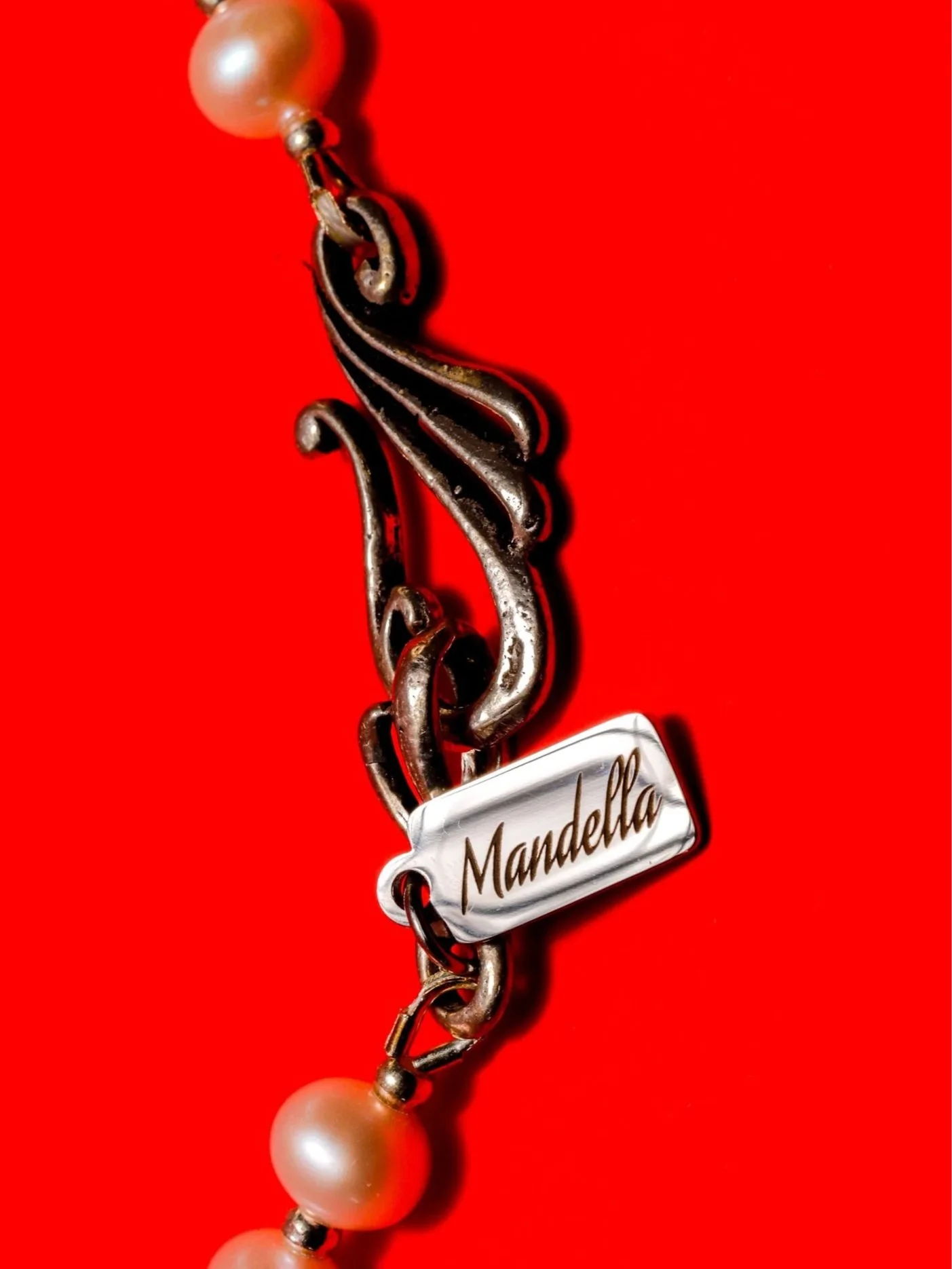 A bracelet with large pearl beads and metal links, featuring a tag with the name "Mandella" on it, against a bright red background.
