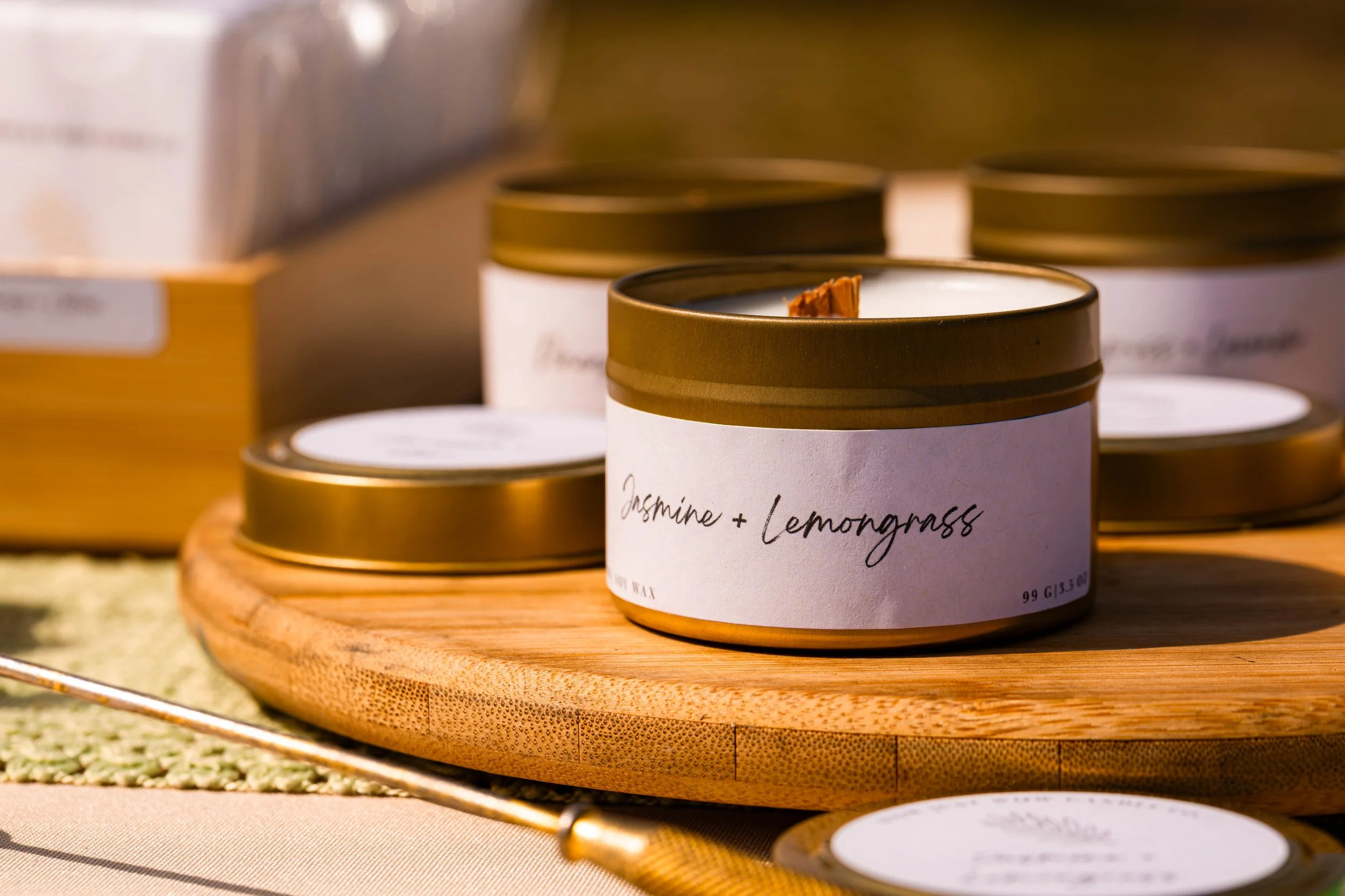 Candle labeled 'Jasmine + Lemongrass' on a wooden round tray, with additional candles and packaging in the background.