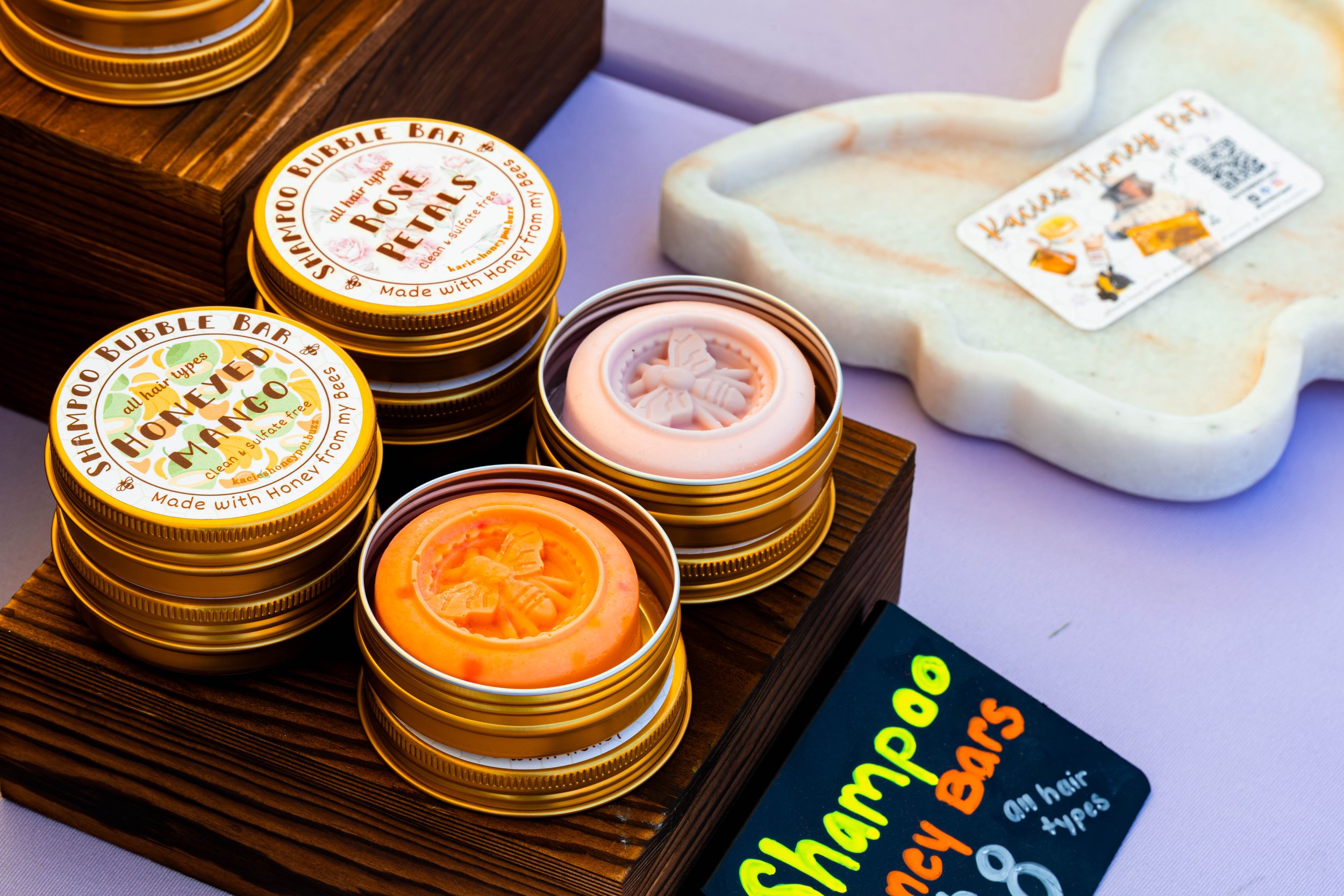 Various tins of hair pomade with bee and honey theme, displayed on a wooden board, including flavors like rose petal, honeyed mango, and honey.