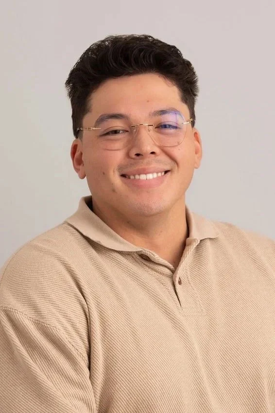 A young man with short dark hair wearing glasses and a beige polo shirt, smiling at the camera against a plain light background.