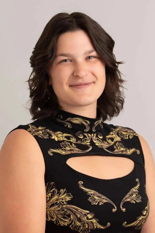 A woman with shoulder-length brown hair and light skin, smiling gently, wearing a sleeveless black top with gold ornate embroidery and a cutout design.