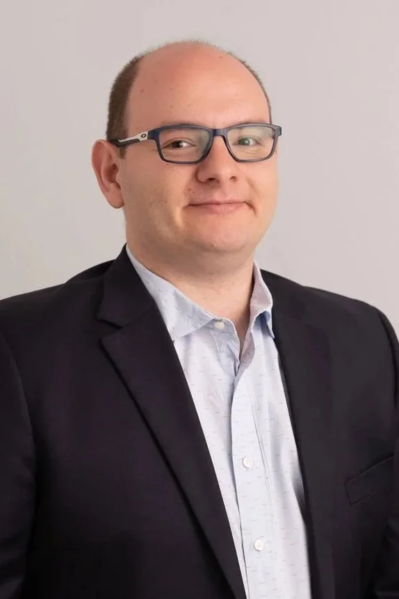 A man with glasses, light skin, and a receding hairline, wearing a black blazer and light-colored dress shirt, standing against a plain light-colored background.