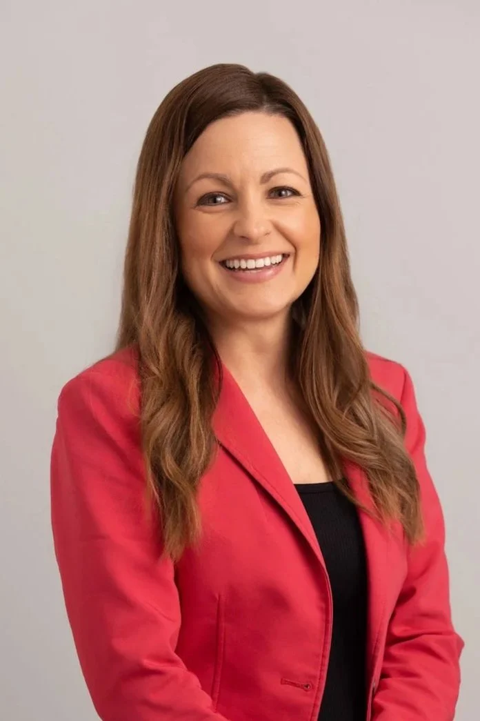 A woman with long brown hair wearing a red blazer and black top, smiling at the camera against a plain light background.