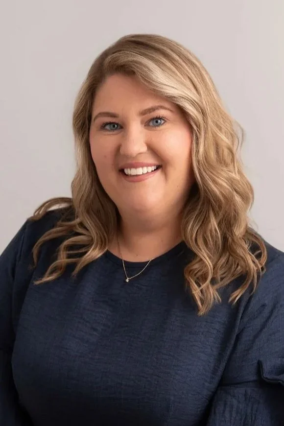 A smiling woman with shoulder-length wavy blonde hair wearing a navy blue top and a delicate necklace.