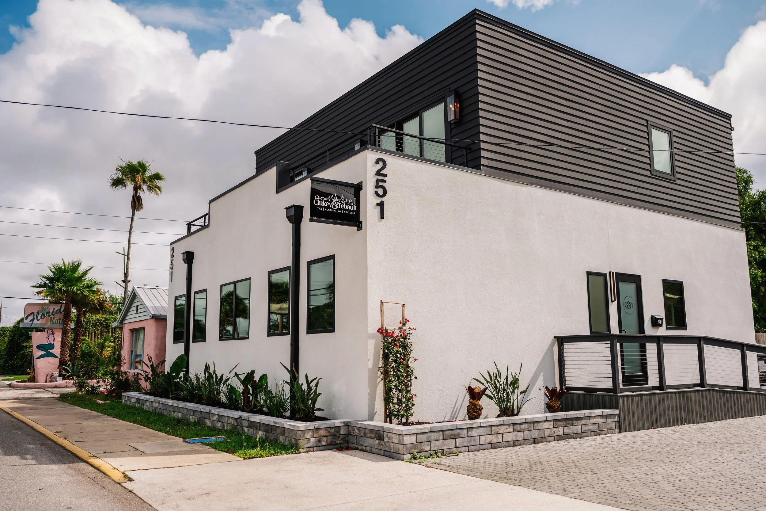 Modern two-story building with black siding and white walls, large windows, a sign for 'Chukey & Rebault Tax Accounting Advising,' street number 251, and landscaped plants in front. Palm trees and cloudy sky in the background.