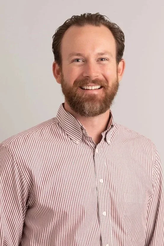 A man with light skin, reddish-brown beard and hair, smiling, wearing a red and white striped button-up shirt, standing against a plain light gray background.