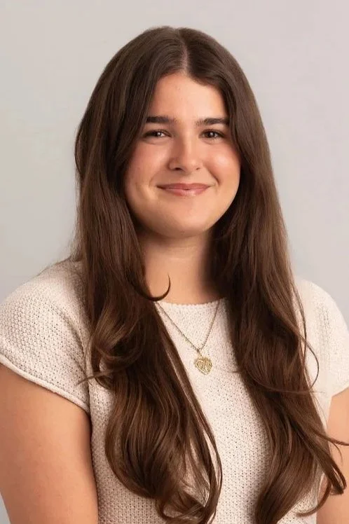 A young woman with long, wavy brown hair, smiling, wearing a light-colored short-sleeve top and a gold heart pendant necklace.
