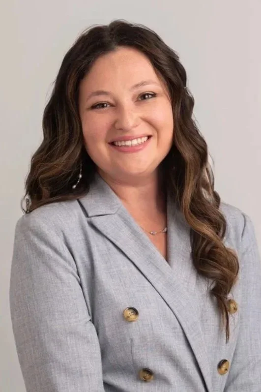 A woman with long wavy brown hair smiling, wearing a light gray blazer with gold buttons against a plain background.