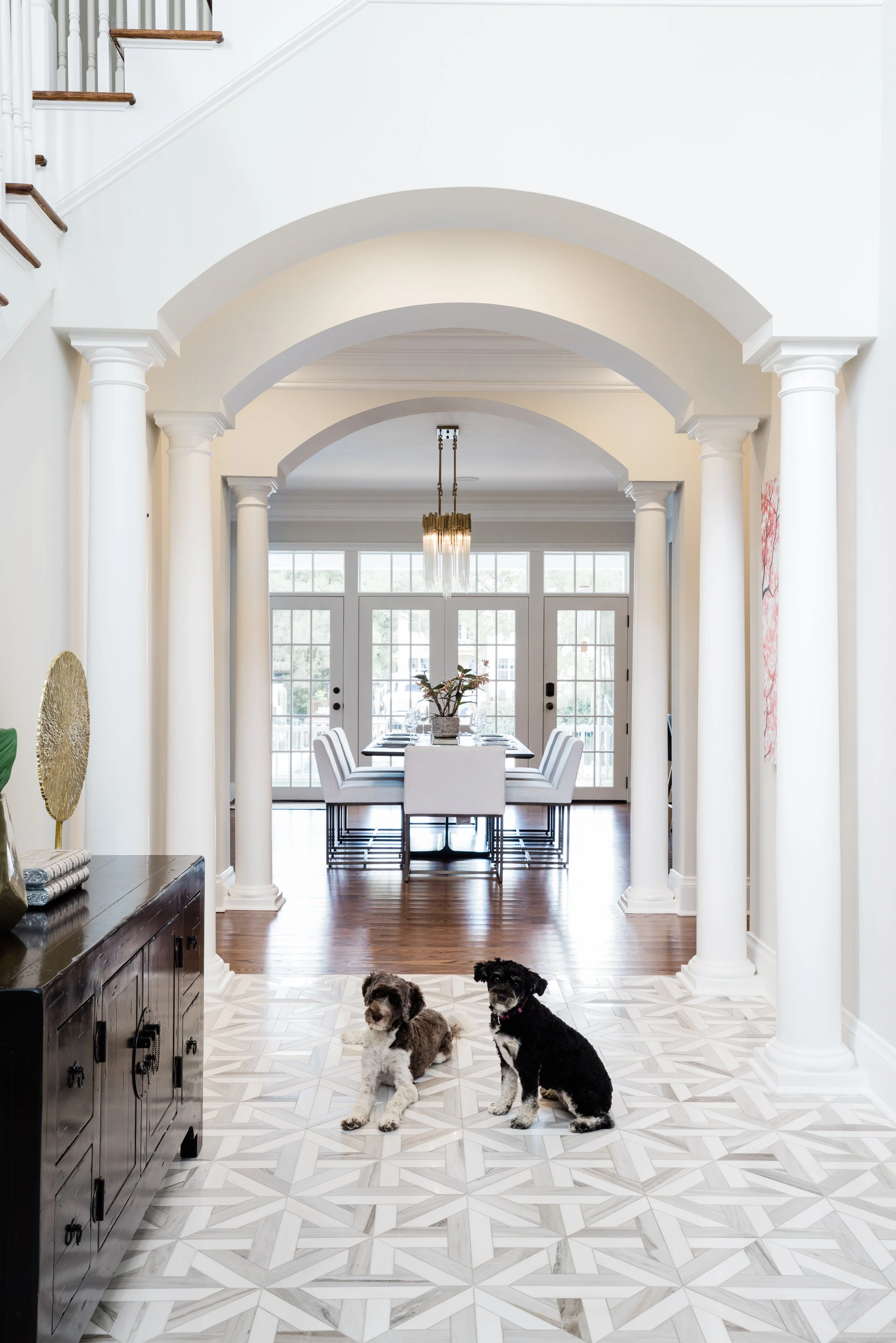 A spacious hall with white columns and arches leading to a dining area. Two dogs are sitting on a patterned tile floor in the foreground. There are large windows and a glass door at the back, with a modern chandelier hanging above the dining table.