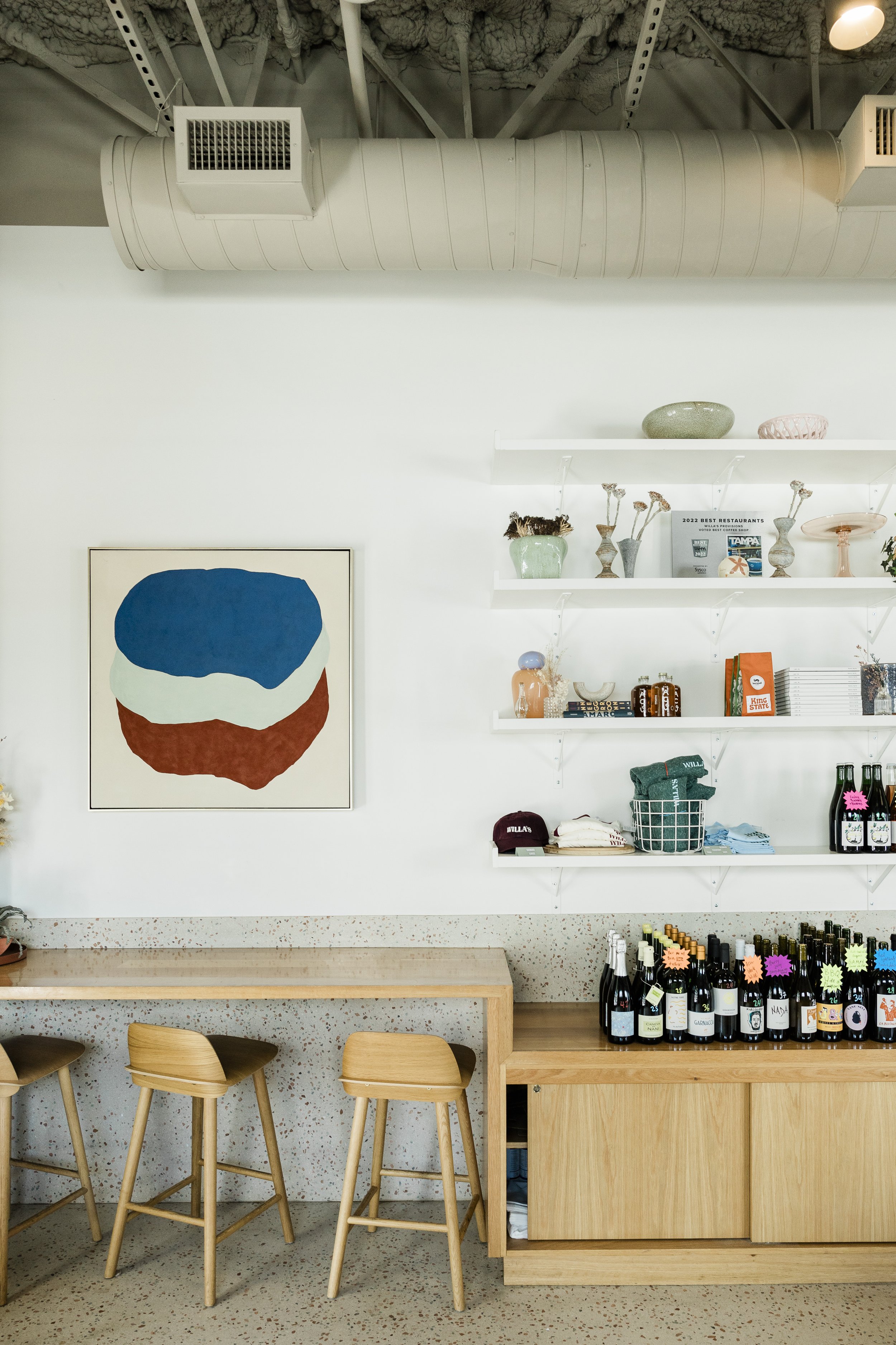 Interior of a modern cafe or shop with three wooden chairs at a counter, a white wall with a framed painting of abstract shapes, floating white shelves holding various decorative items like vases and plants, and a display of bottled beverages with co