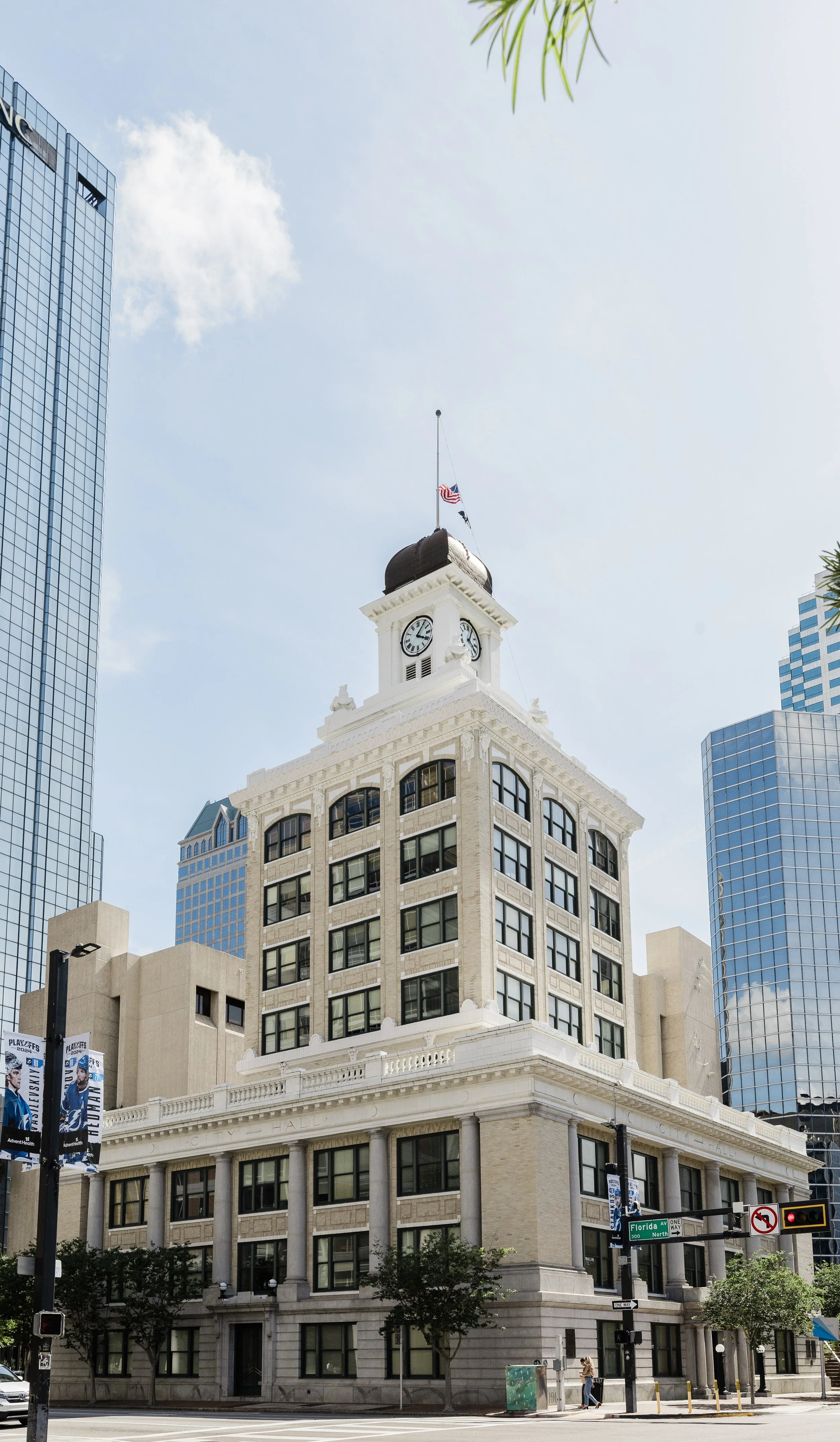 A historic white clock tower building with a brown rooftop, surrounded by modern glass skyscrapers in an urban city scene on a clear day.