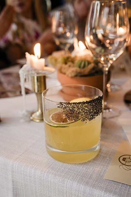A glass of yellow cocktail with a lime slice and black sugar rim on a table set for a fancy dinner, with candles and wine glasses in the background.