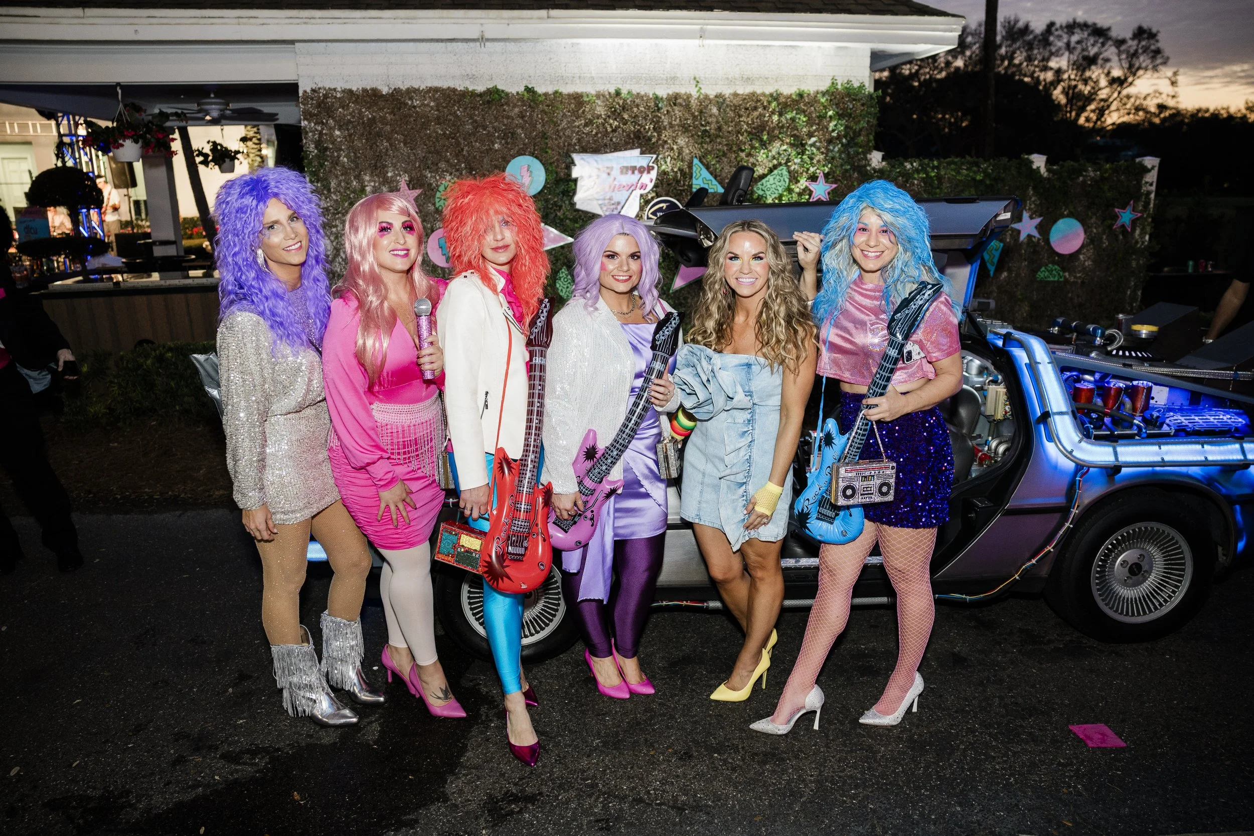 Seven women dressed in colorful 80s style costumes, standing together outdoors at sunset, with a DeLorean car behind them decorated with neon signs and stars.