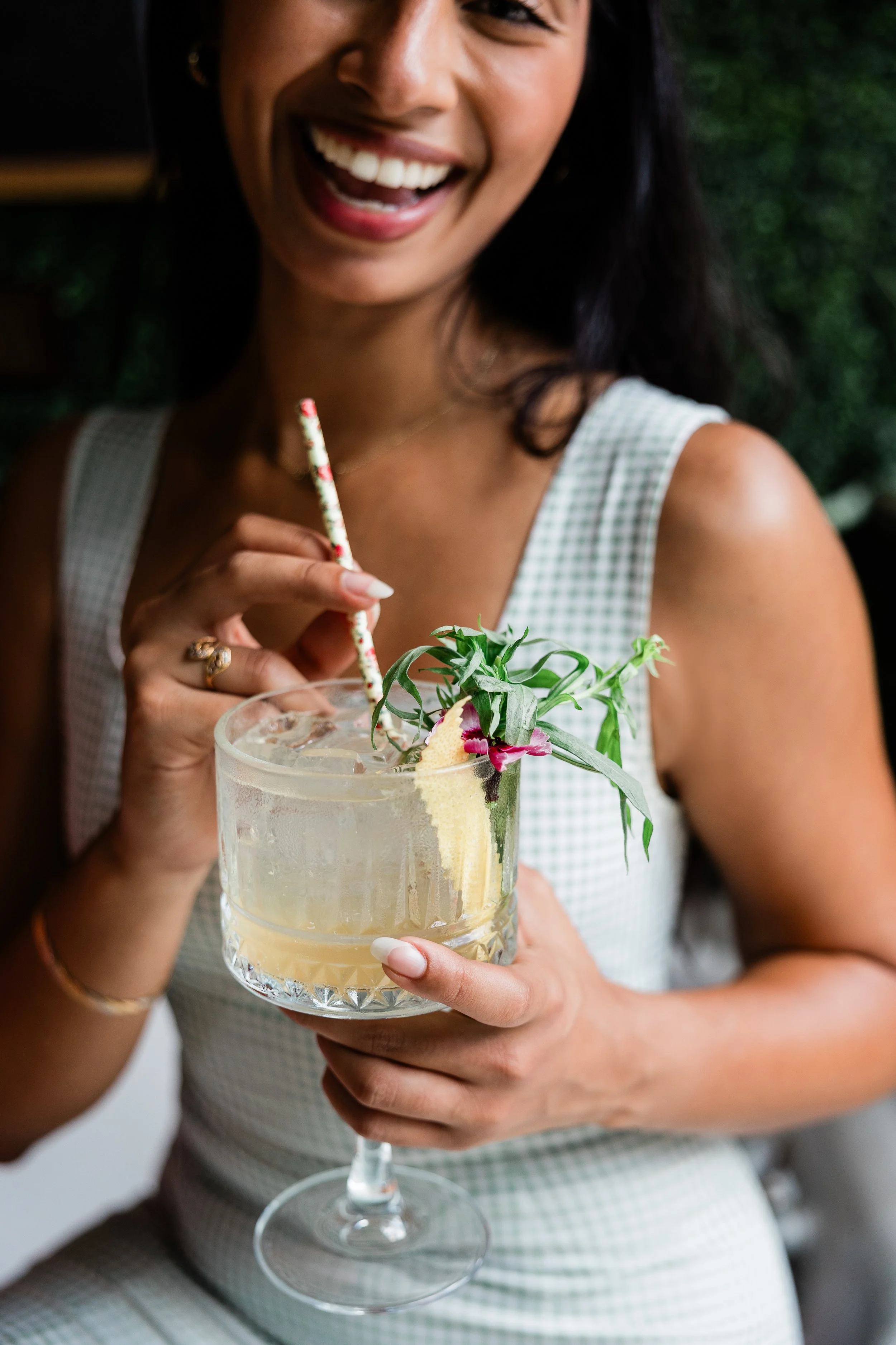 A woman with dark hair and a checkered dress smiling while holding a large glass of cocktail with flowers and a decorative straw.