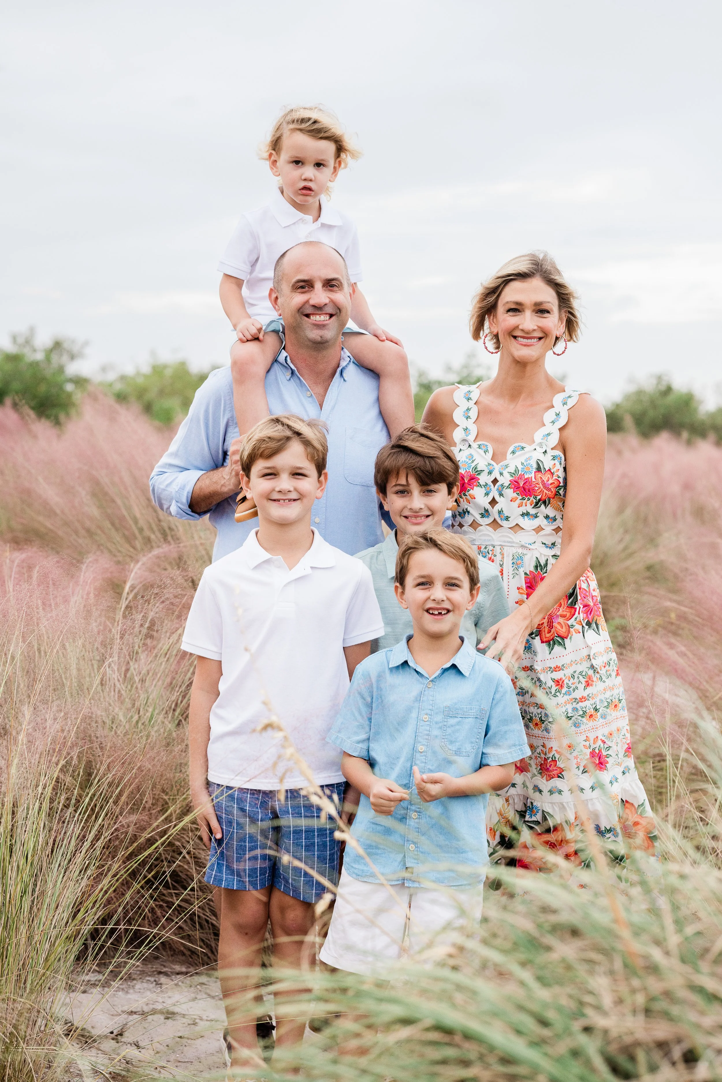 A happy family of seven standing outdoors in a grassy field with a cloudy sky, including a man carrying a young boy on his shoulders, a smiling woman, and four children, some of whom are smiling.