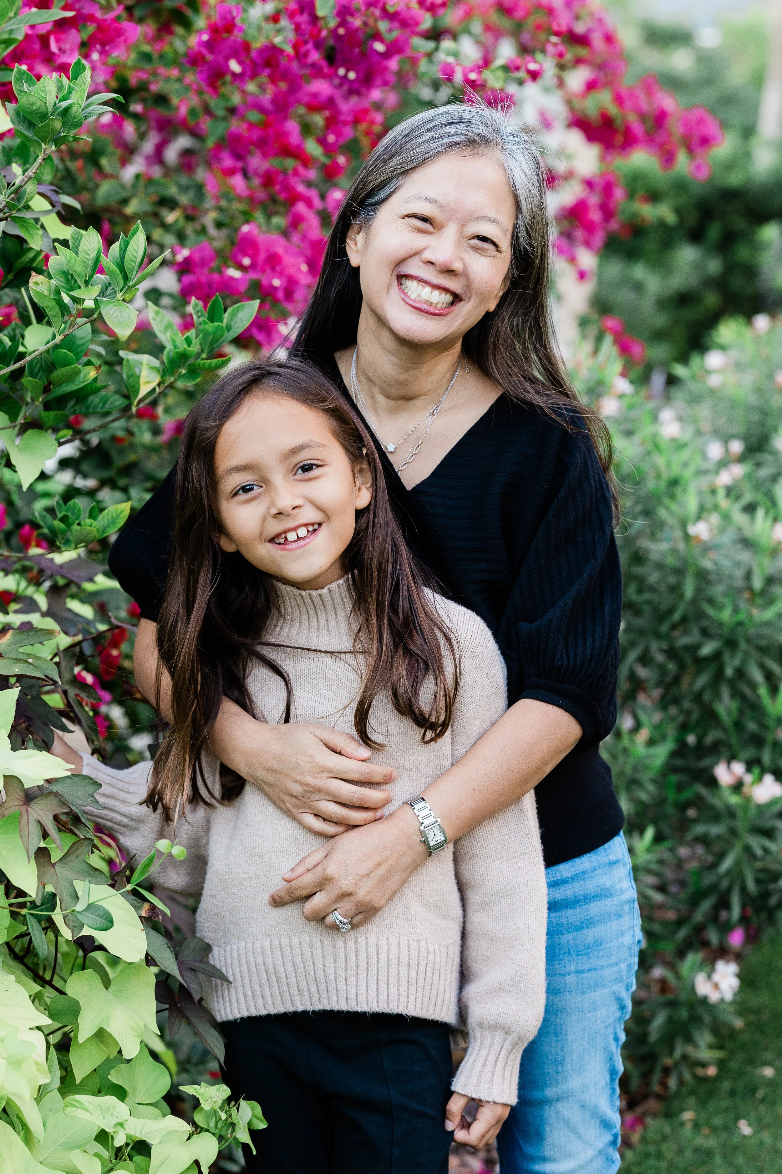 A woman and a young girl standing outdoors in front of pink and purple flowers, smiling at the camera.