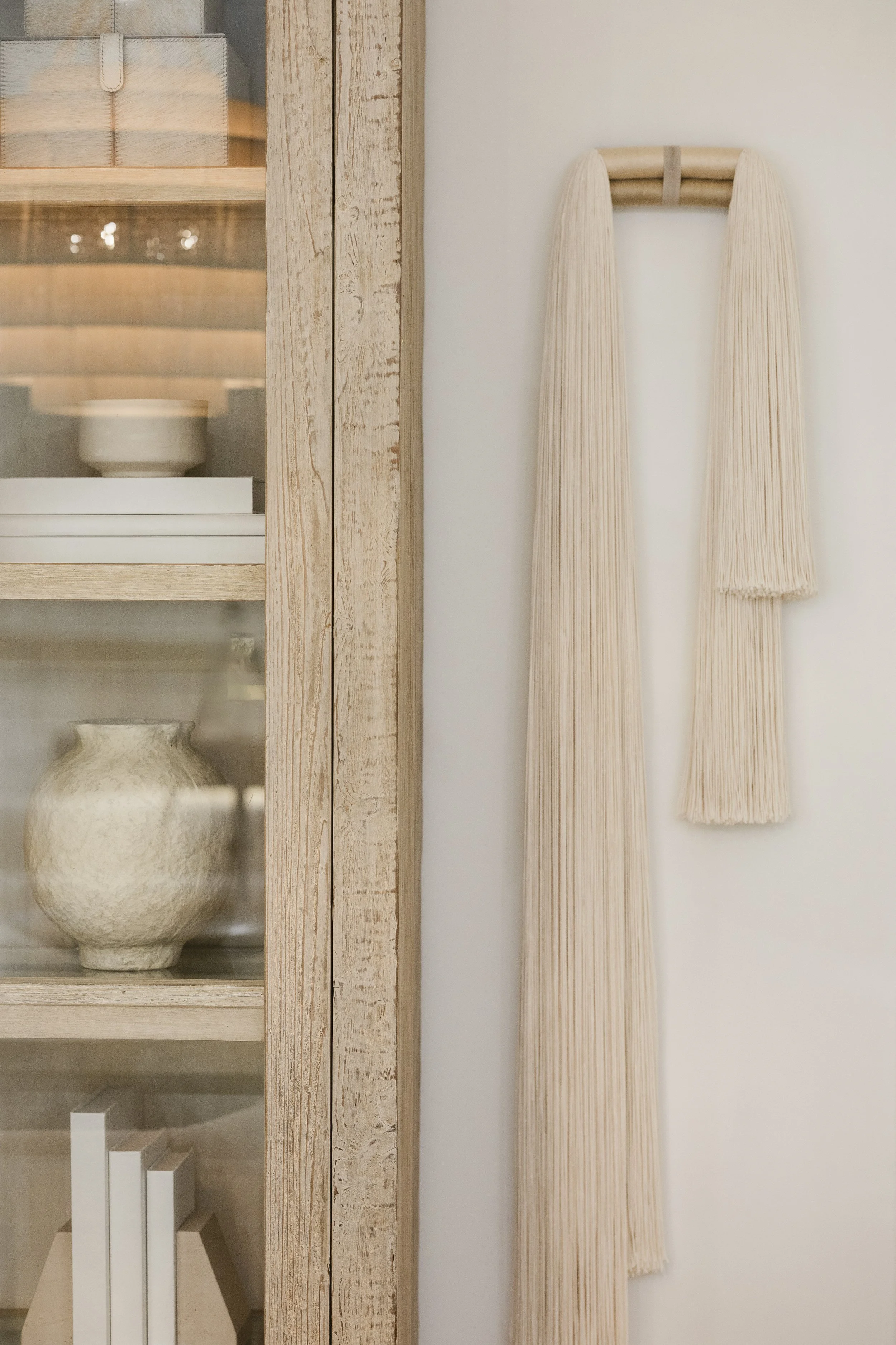 A beige wall decor piece shaped like a long scarf with fringe, mounted on the wall, next to a wooden cabinet with glass doors.