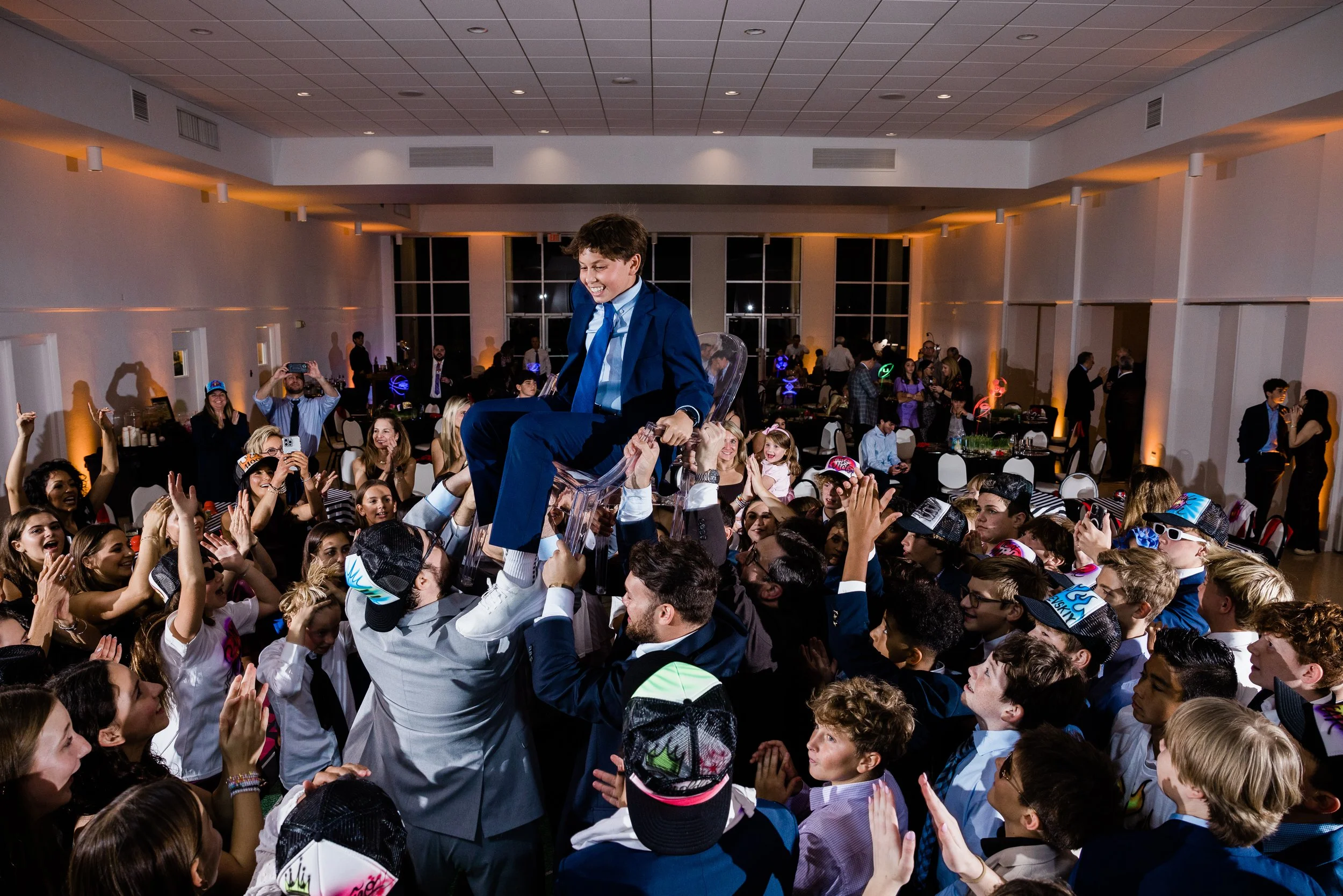 Crowd of people at a party lifting a young boy in a blue suit on a clear acrylic chair. The boy is smiling and appears to be happy. The room is decorated with orange lighting and there are several people taking photos and enjoying the event.