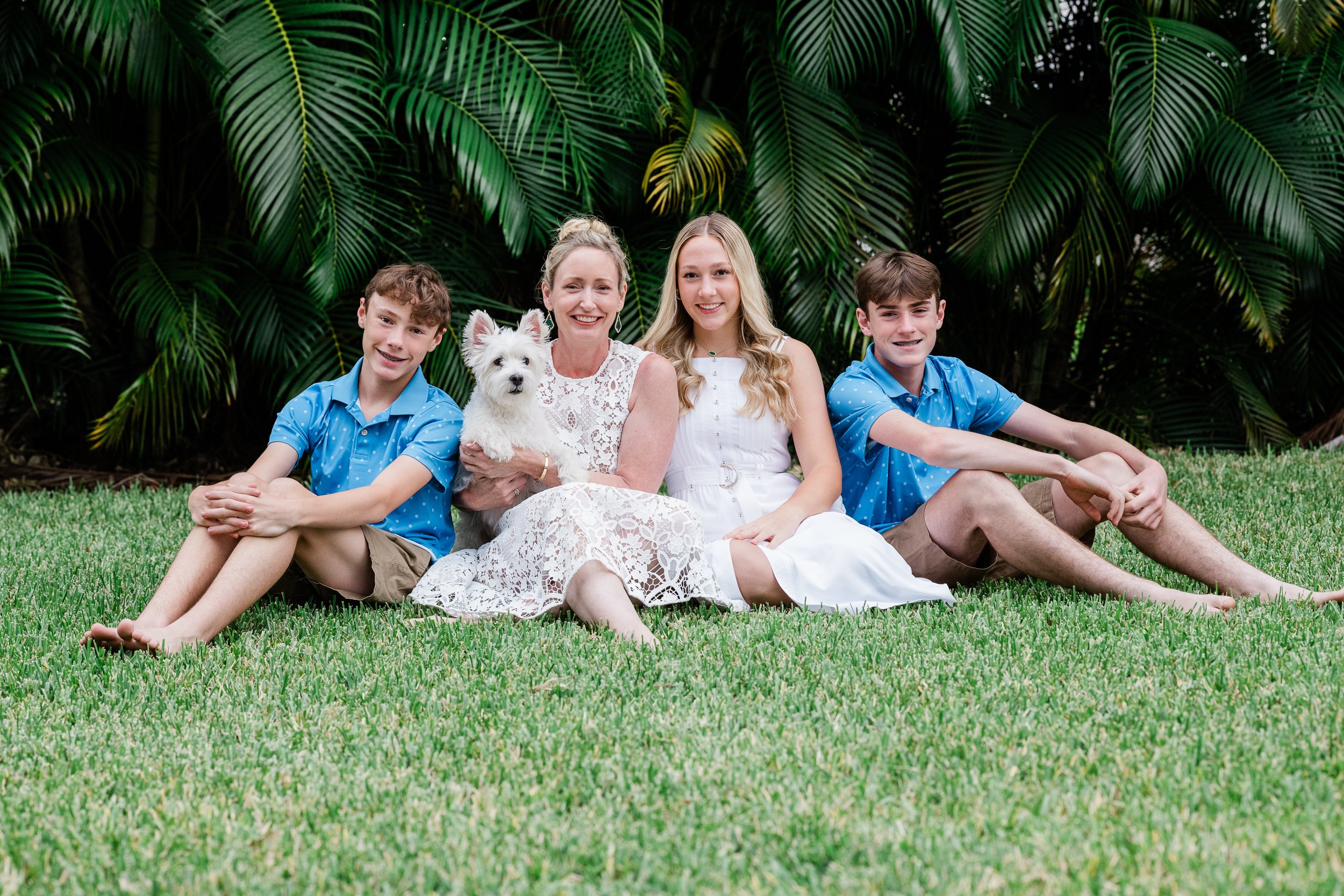 A family of five, including an older woman, two teenage girls, two teenage boys, and a white dog, sitting together on grass in front of large tropical plants.