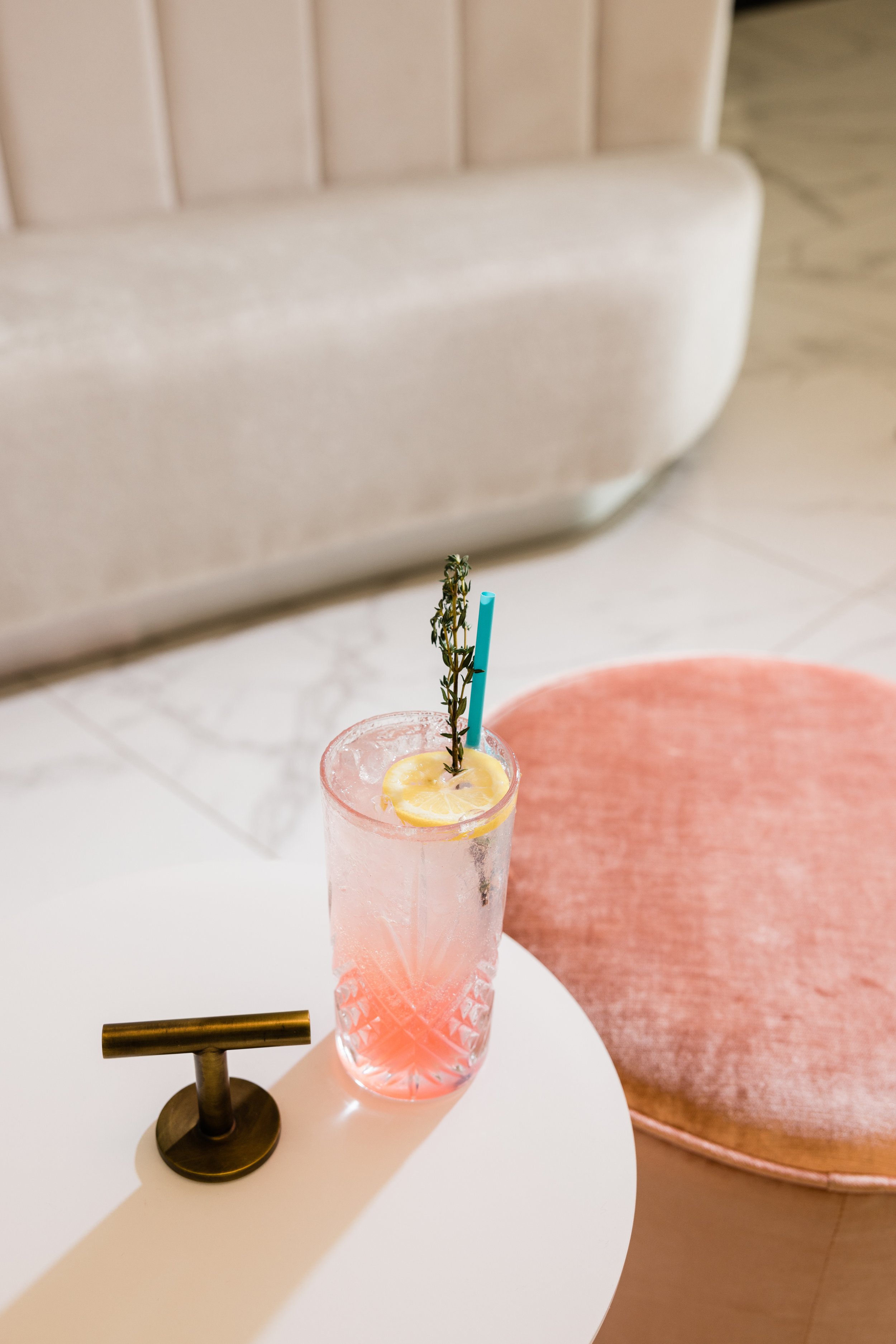 A pink cocktail in a tall glass with a lemon slice, a sprig of thyme, and a blue straw, placed on a white table near a brass-colored table lamp, with a beige upholstered chair in the background.