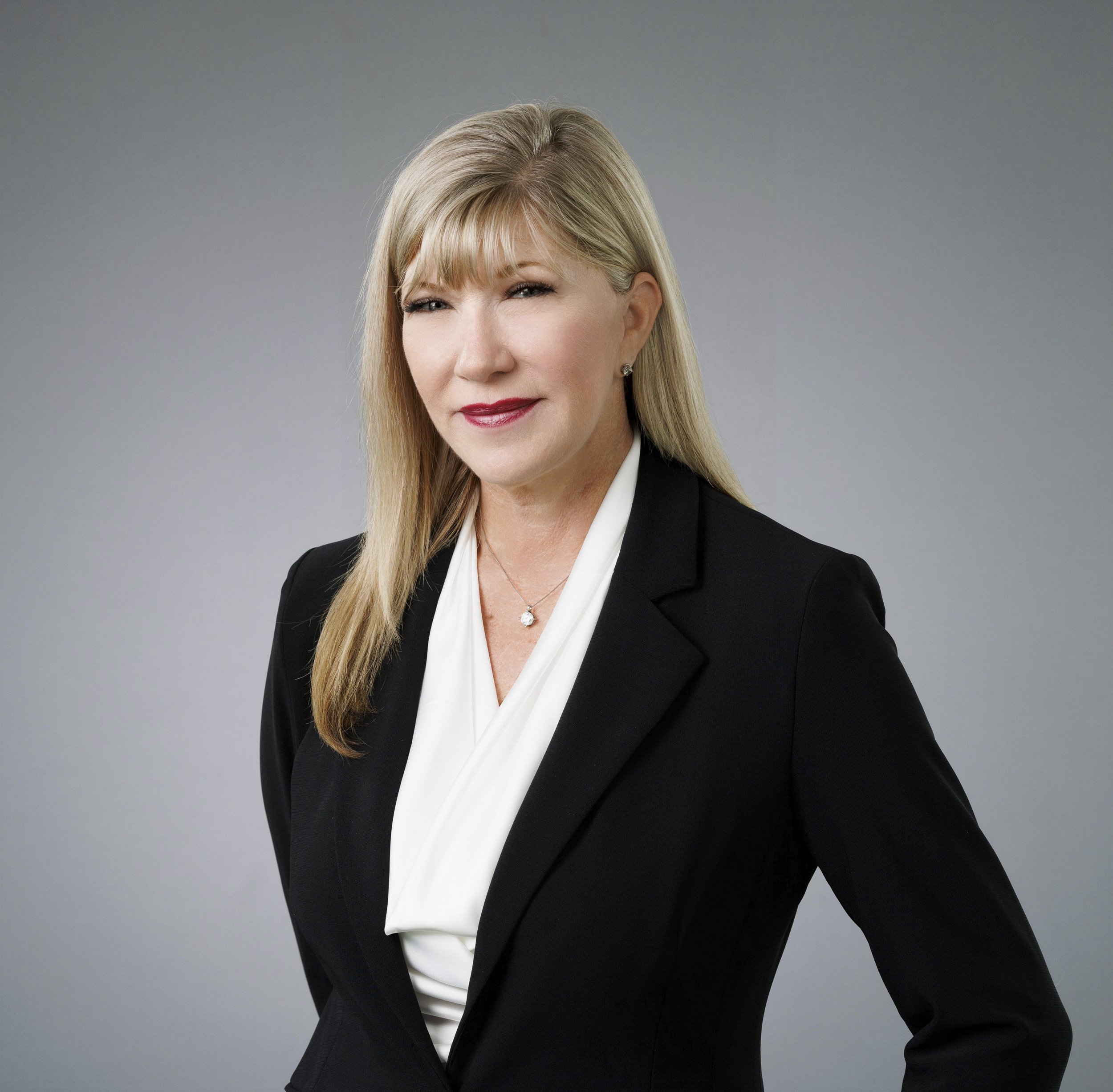Close-up of a middle-aged woman with blonde hair wearing a black blazer and a white blouse, smiling slightly, against a gray background.