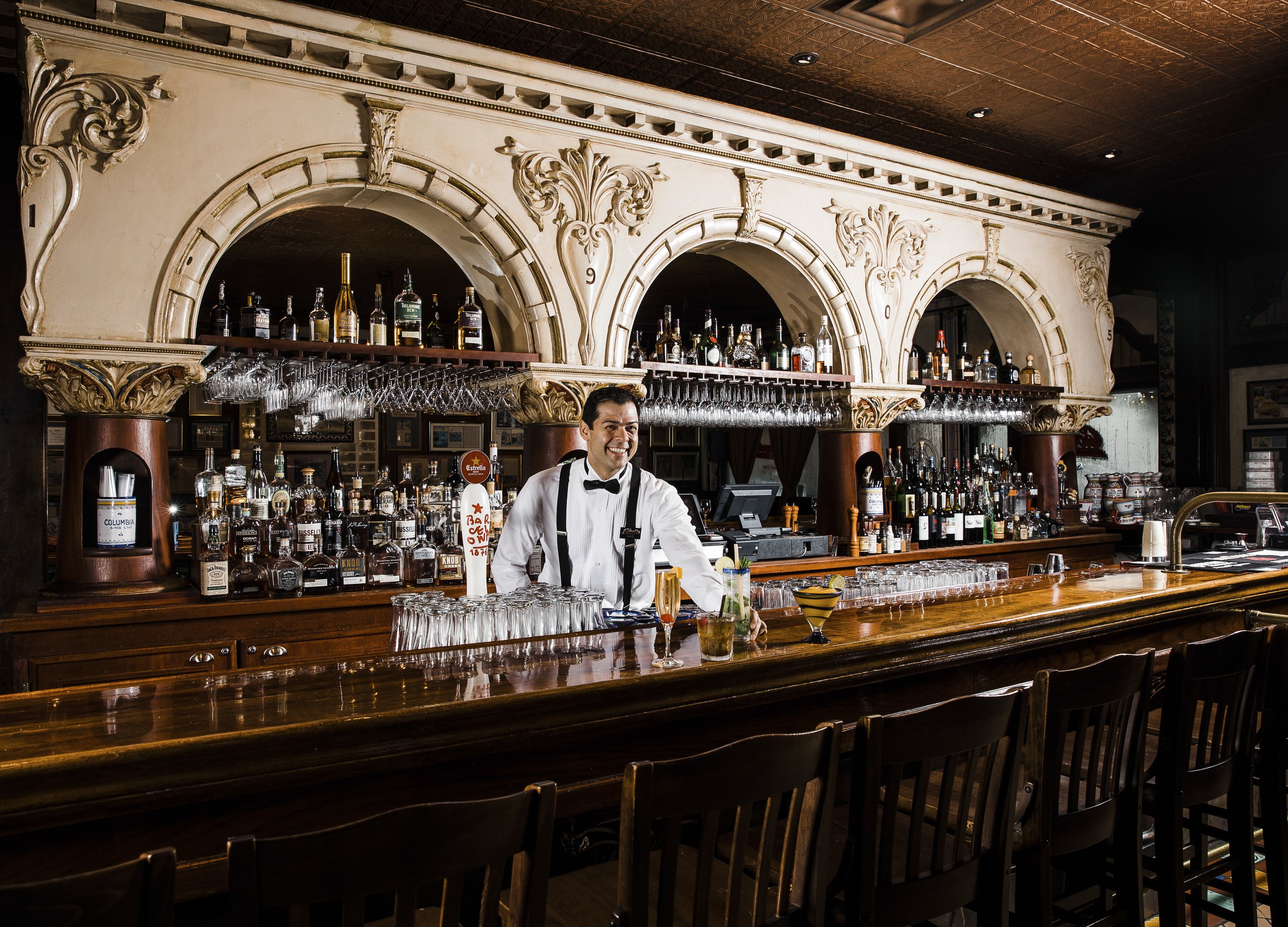 Bar of Columbia Restaurant with ornate decor, glass bottles of alcohol on shelves, and a smiling bartender in a white shirt and black bowtie.