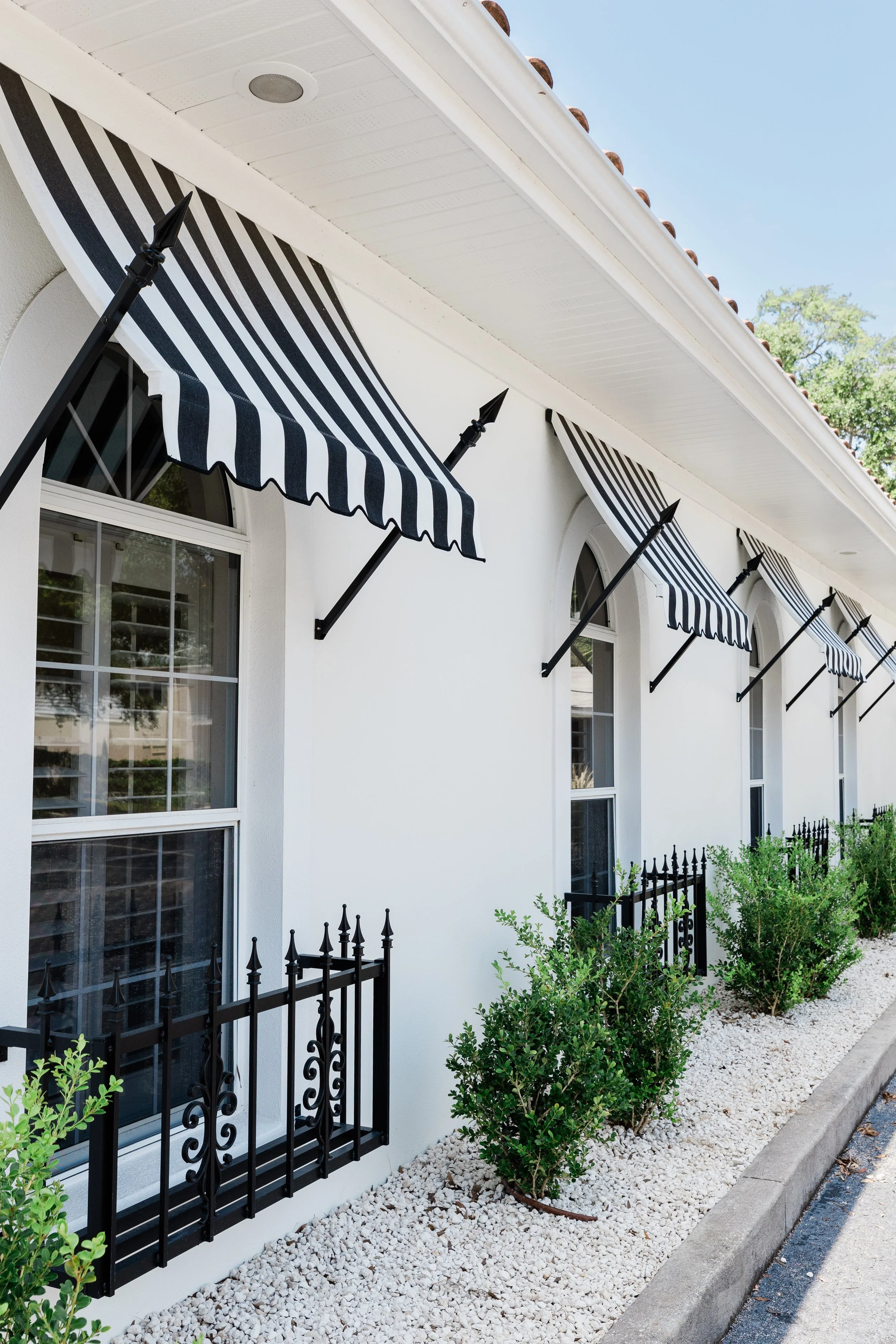A row of windows with black and white striped awnings on a white building. The area below the windows is landscaped with small green shrubs and white gravel, bordered by a concrete sidewalk.