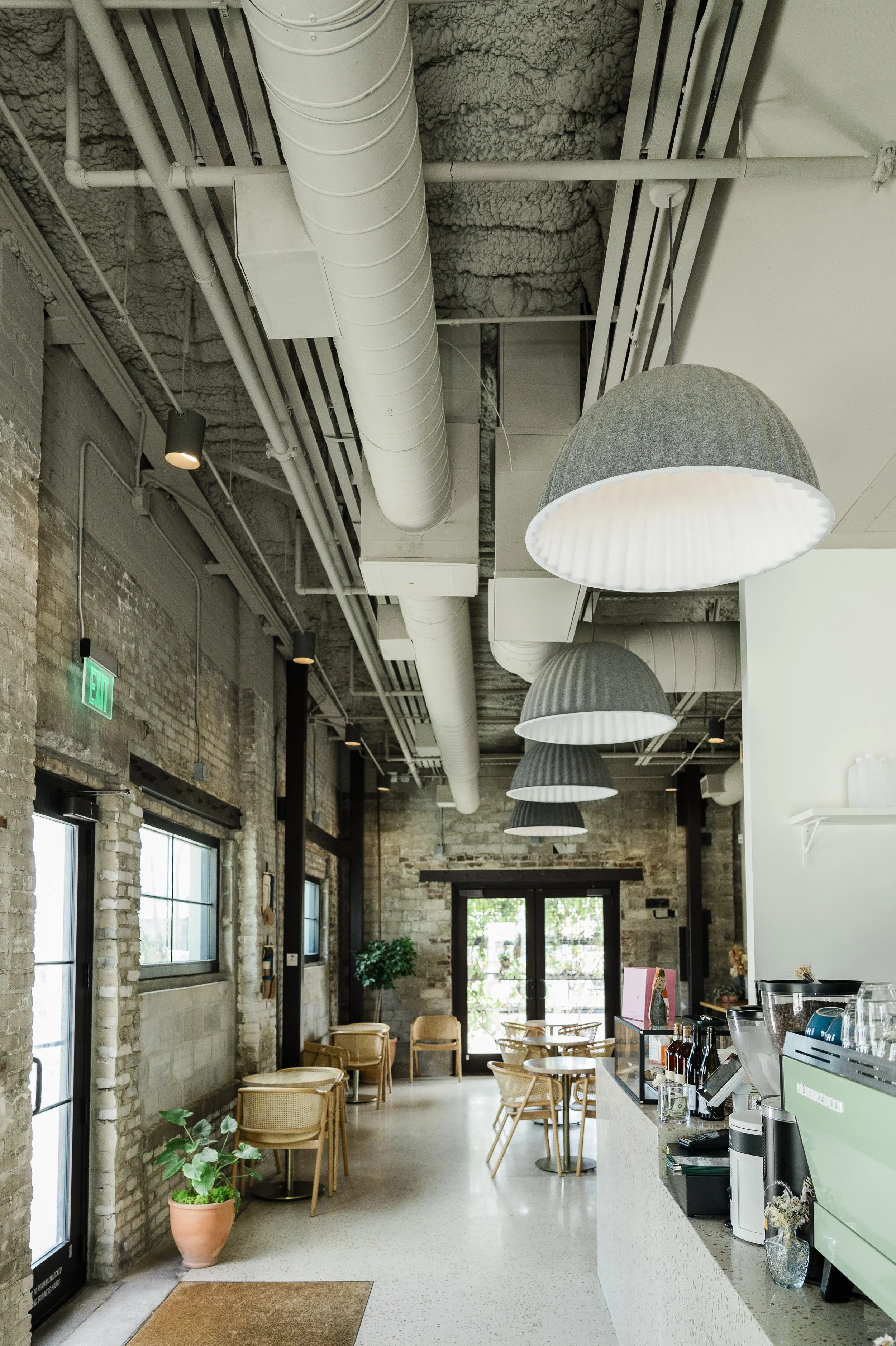 Interior of a coffee shop or cafe with exposed brick walls, overhead industrial piping, hanging black and white metal light fixtures, and wooden chairs around small tables near large windows.