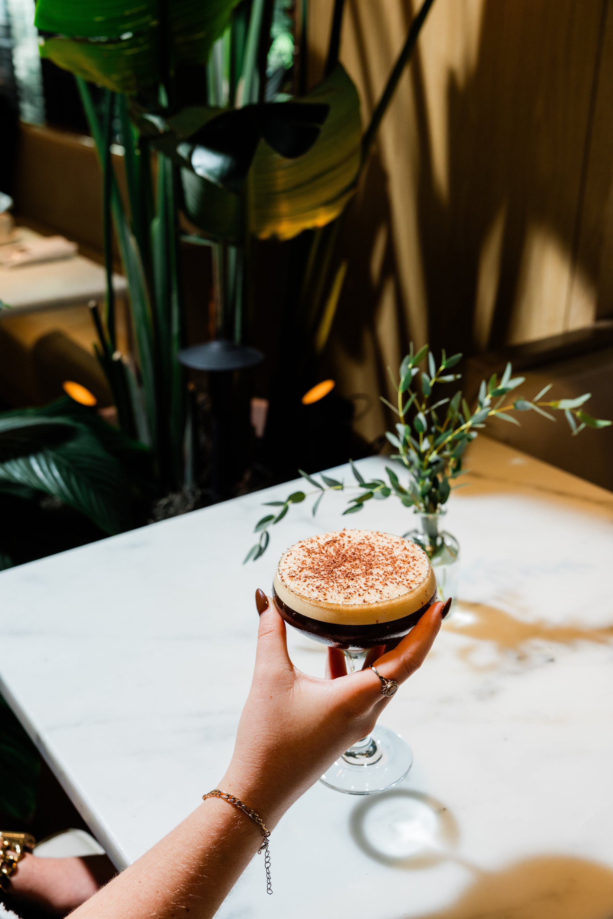 A person holding a coffee or dessert drink in a glass with a frothy top sprinkled with cocoa powder, with a background of green plants and a wooden wall, on a white marble table.