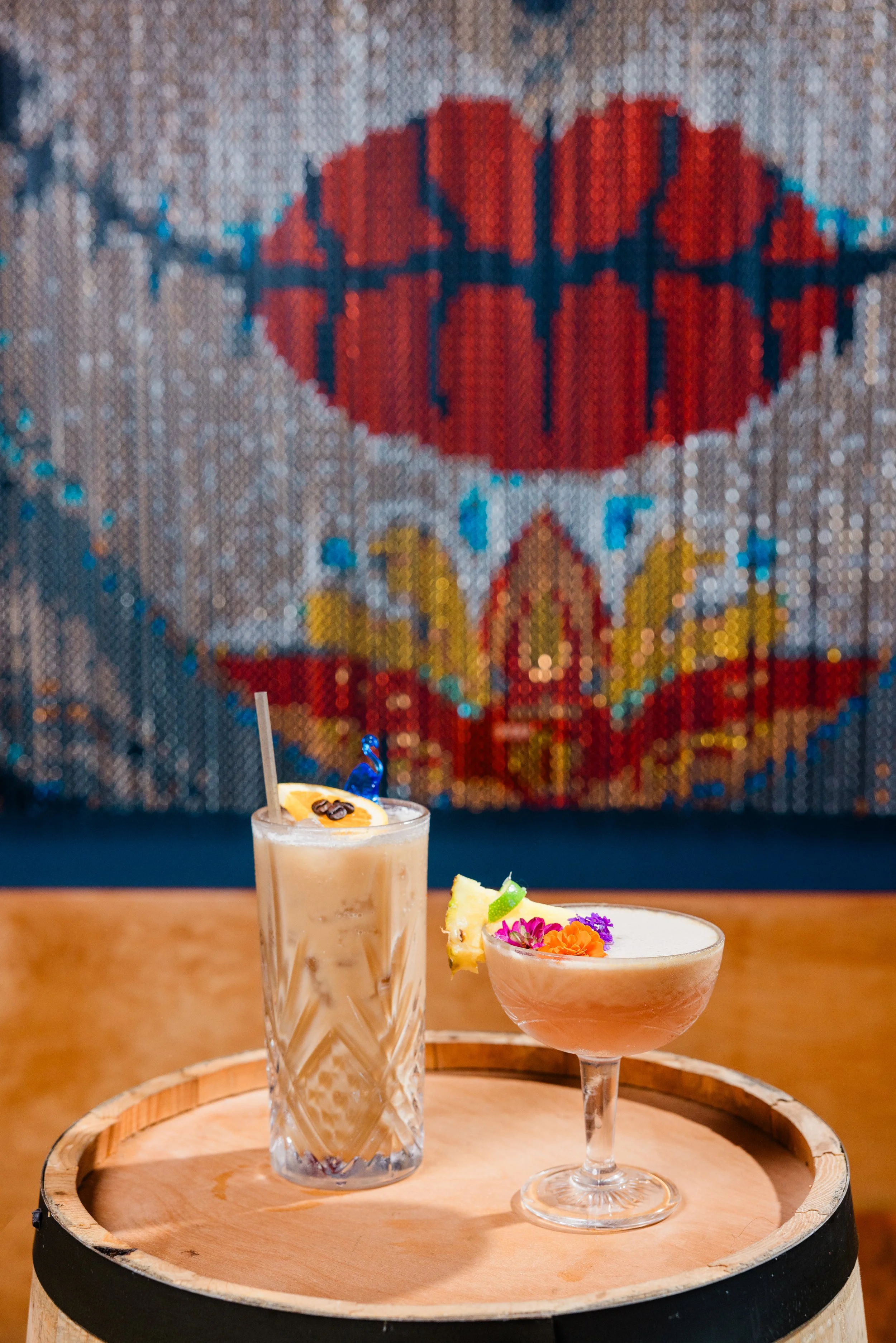 Two drinks on a round wooden tray in front of a colorful mosaic heart artwork.