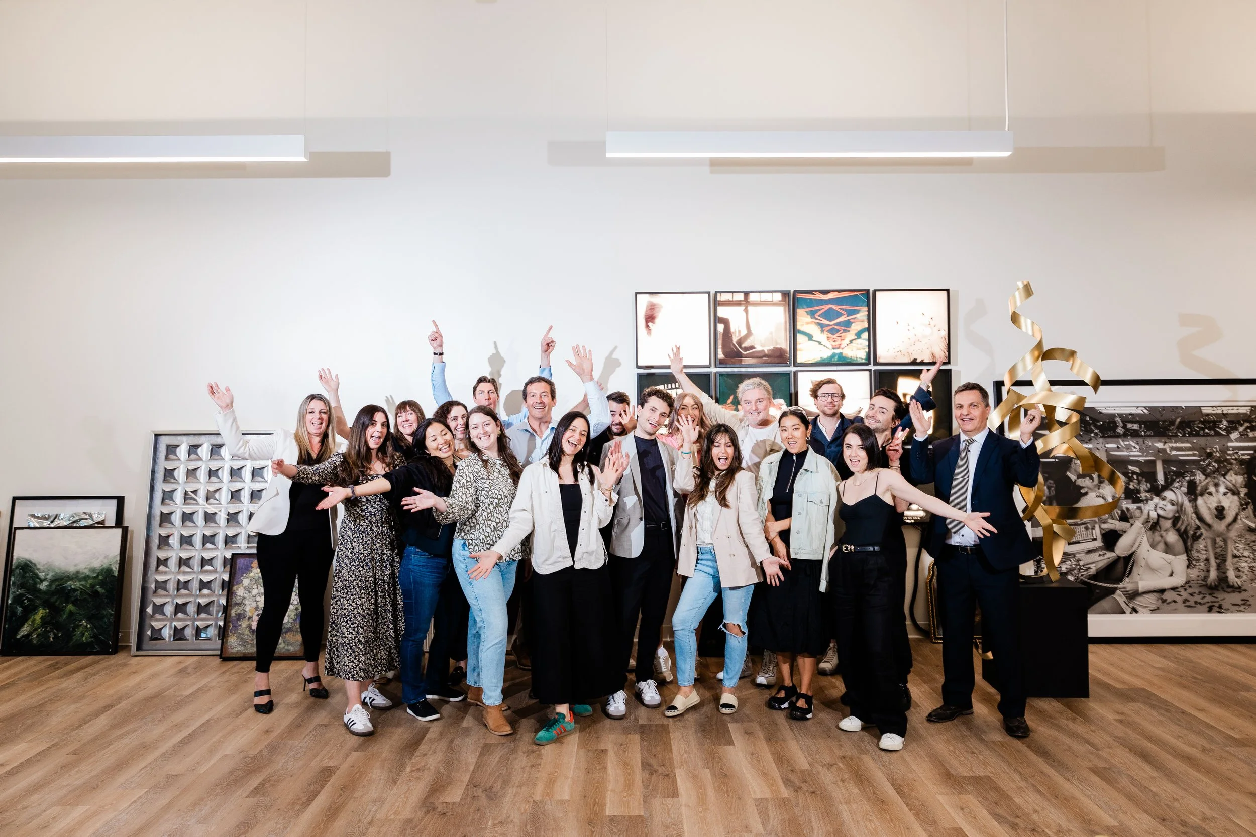 A large group of diverse people celebrating indoors at an art gallery, smiling, with some raising their hands and arms, surrounded by various artworks and photographs on the walls.