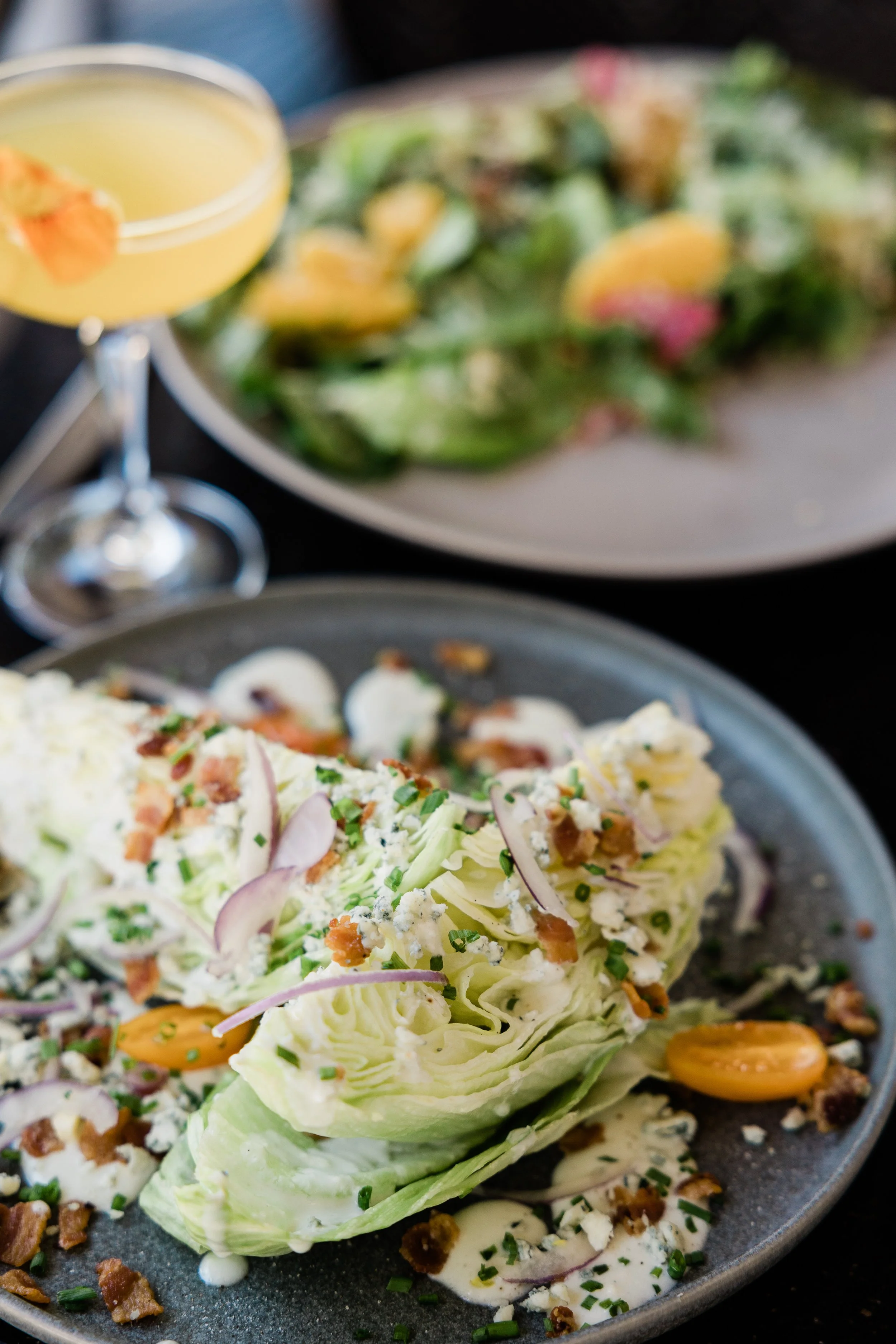 A plate with a wedge of iceberg lettuce topped with blue cheese dressing, bacon bits, chopped chives, and red onion slices. In the background, a cocktail glass with a yellow drink and an orange garnish is visible, along with a blurred salad on a silv